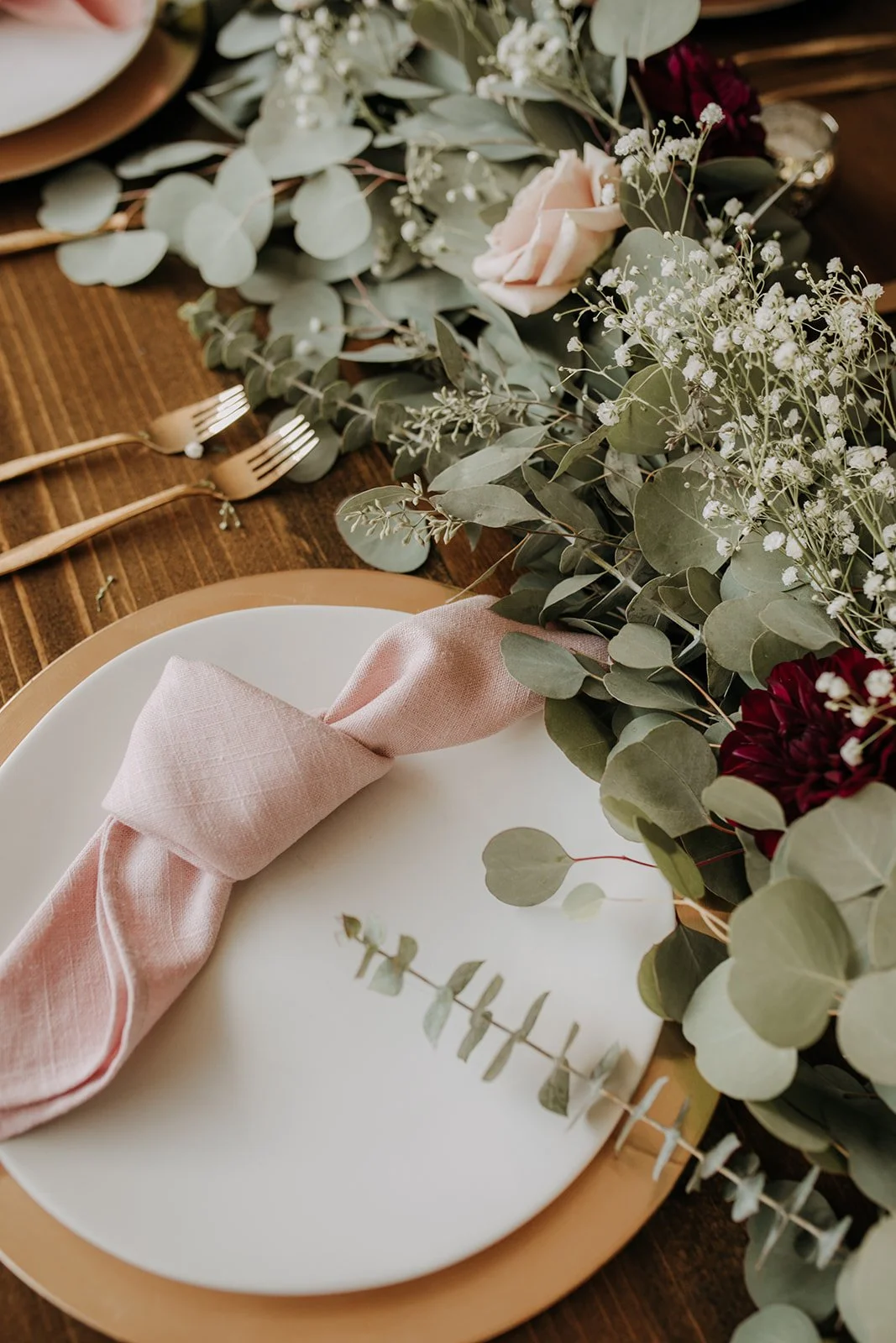 A table setting with a white plate, pink cloth napkin, and floral centerpiece including greenery and flowers.