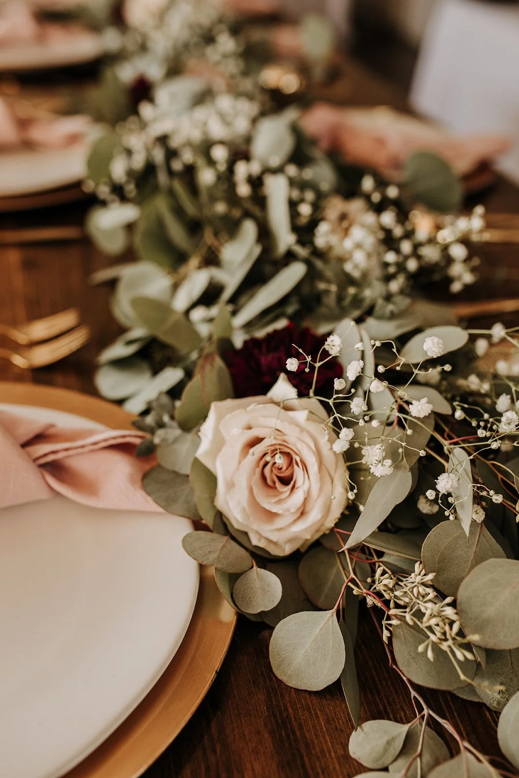 Elegant floral centerpiece with white rose, baby's breath, and eucalyptus on a wooden table with a white plate and blush-colored napkin.