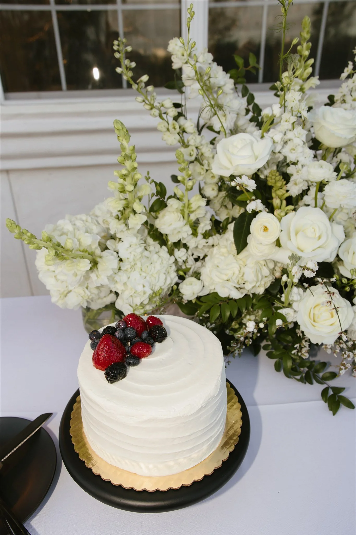 White wedding cake with fresh berries on top placed on a black cake stand, set on a table with a large white floral arrangement in the background.