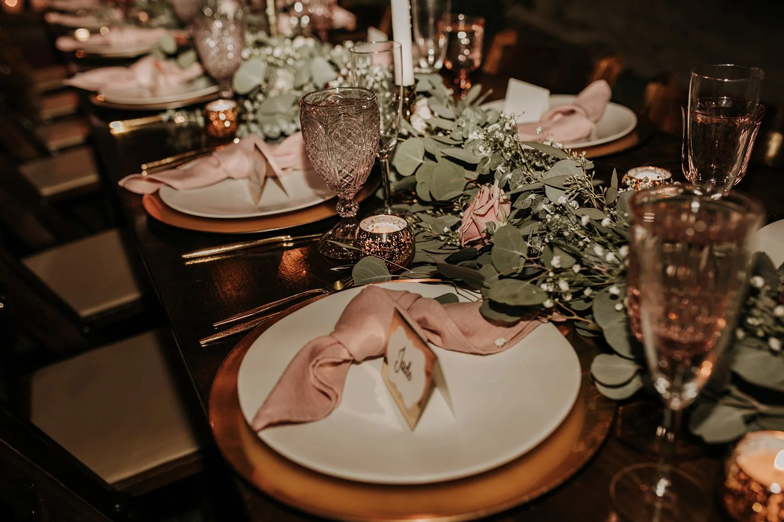 A table set for a formal dinner with white plates, pink cloth napkins, gold utensils, wine glasses, and floral centerpieces with greenery, candles, and pink roses.