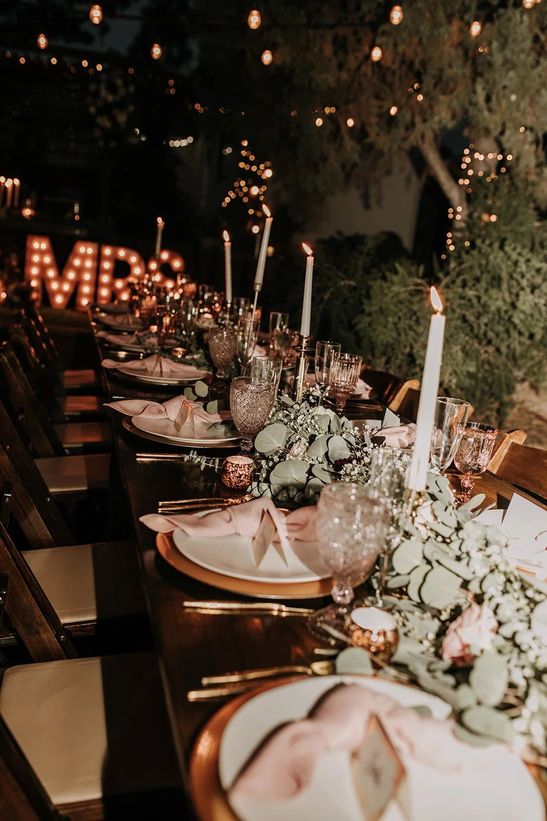 Decorated outdoor dinner table at night with candles, greenery, and pink napkins, featuring illuminated 'MR' and 'MRS' signs in the background.