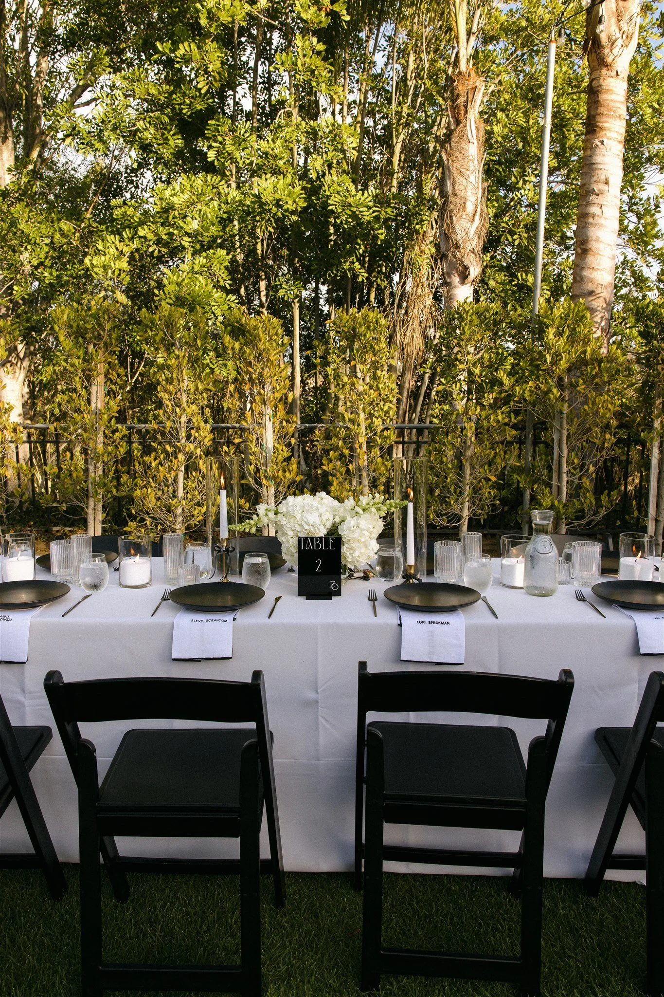 A beautifully set outdoor dining table with black plates, silverware, white napkins, and candles, surrounded by greenery and trees.