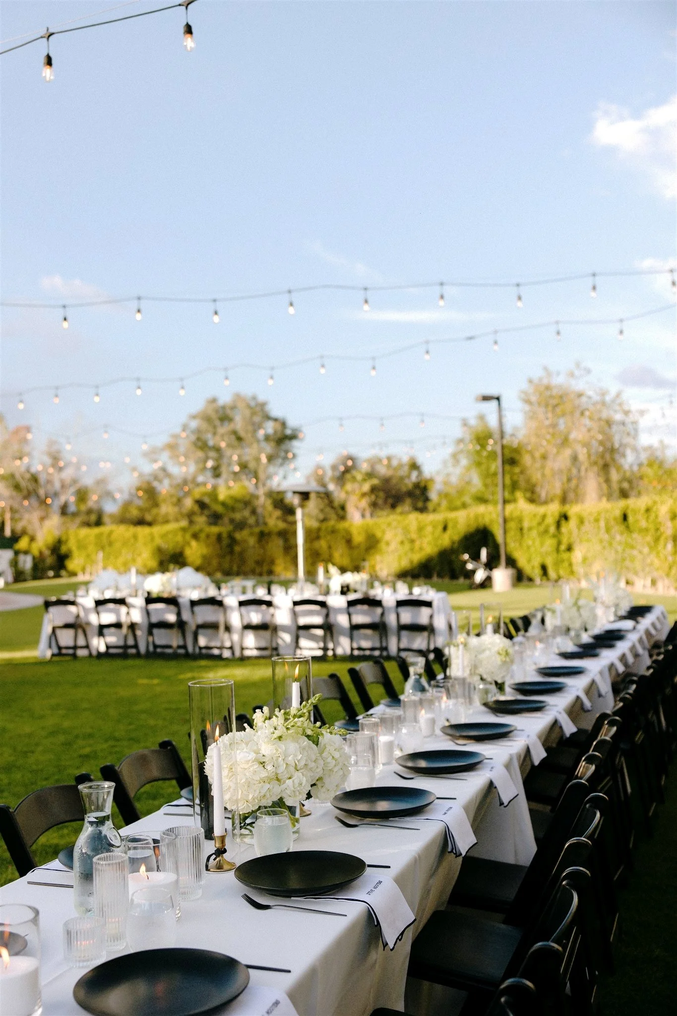 Outdoor banquet table set with black plates, white napkins, tall floral centerpieces, and candles, under string lights, on a green lawn with trees and a clear sky in the background.