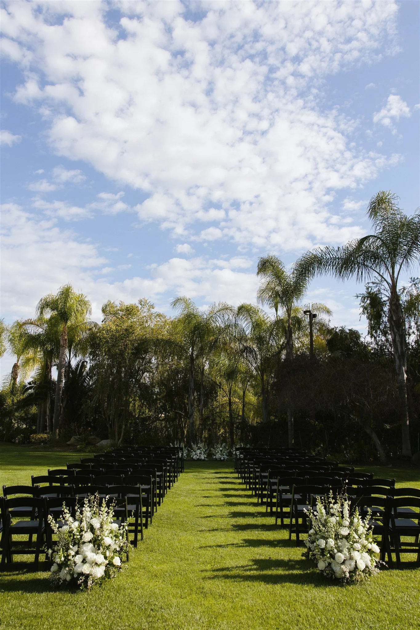 Outdoor wedding ceremony setup with black chairs, white floral arrangements, green grass, tall trees, and partly cloudy sky.
