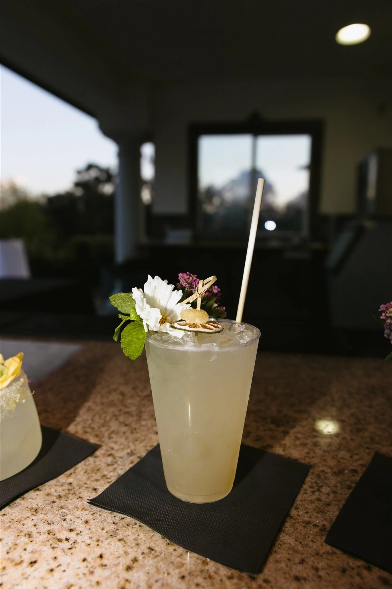 A cocktail with flowers and garnishes on a black napkin, placed on a granite countertop inside a room with a large window and trees outside.