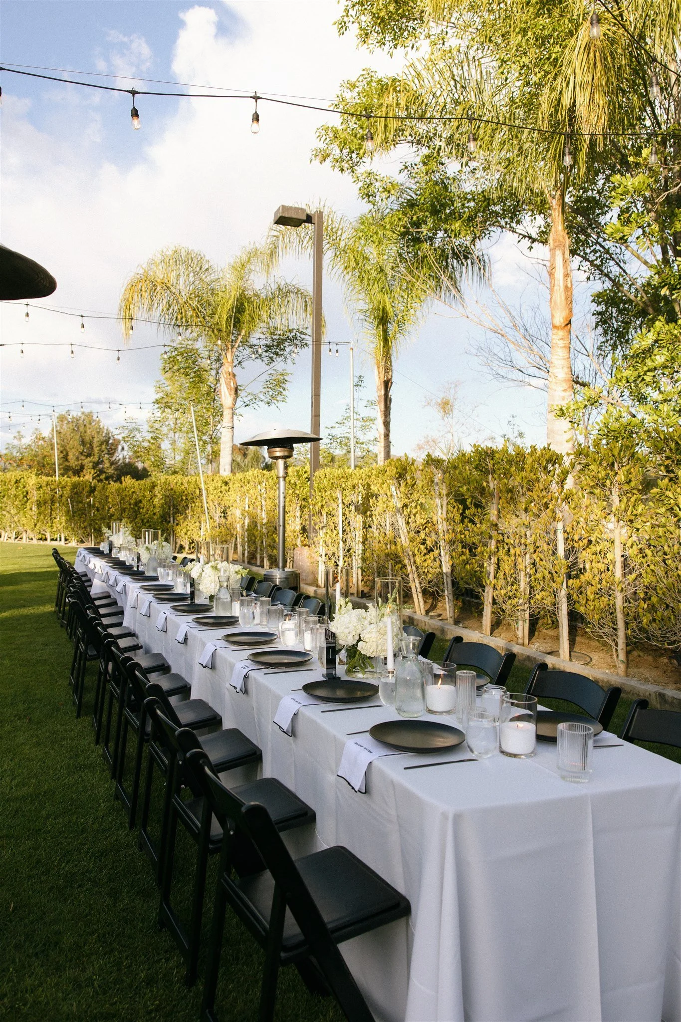 Outdoor dining table set for a large gathering with black plates, white napkins, water pitchers, candles, and floral centerpieces, under string lights and palm trees.