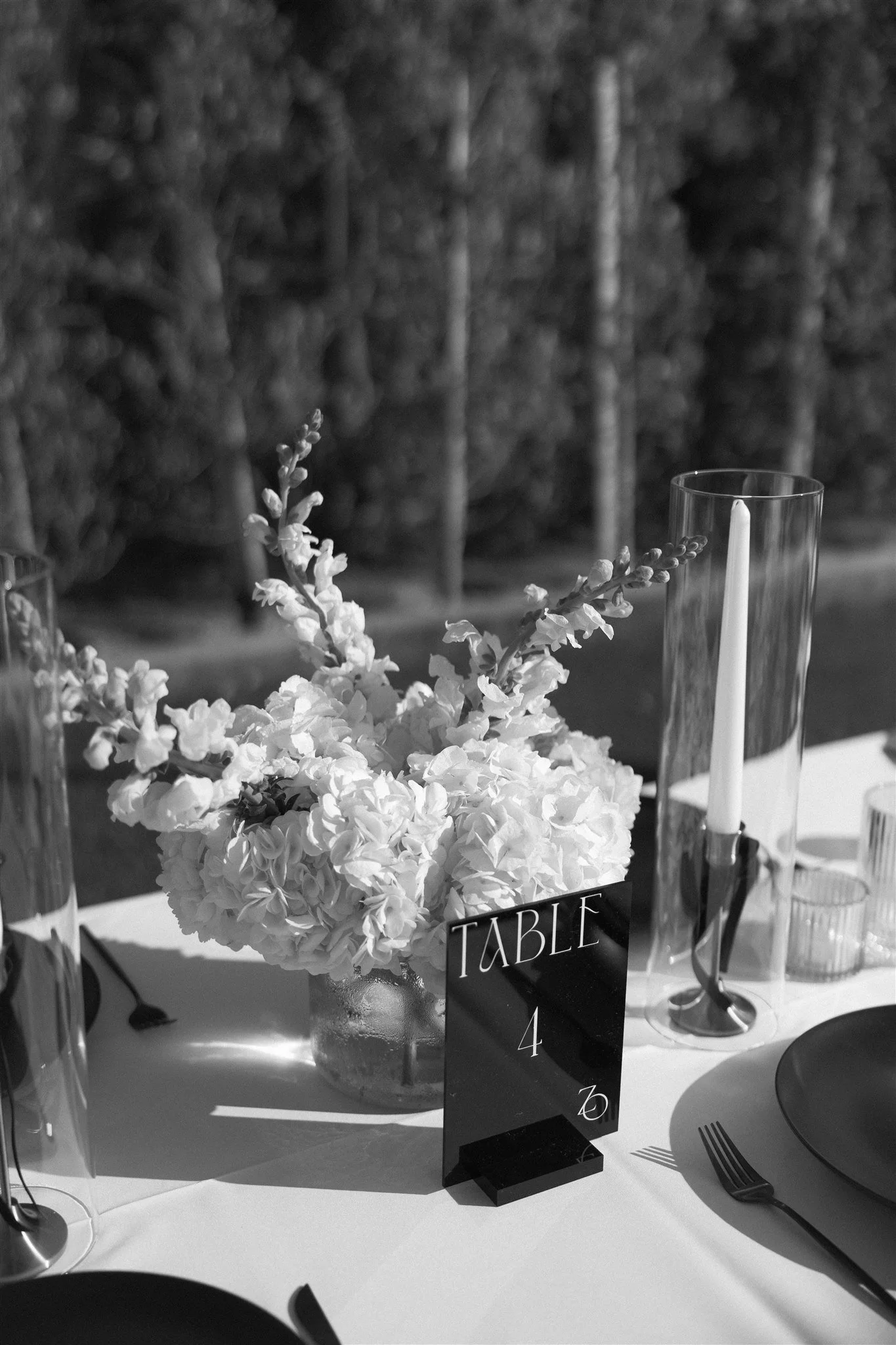 A table centerpiece with a bouquet of white flowers in a vase, a black table number sign for table 4, and several tall glass candle holders with candles inside, set outdoors with trees in the background.
