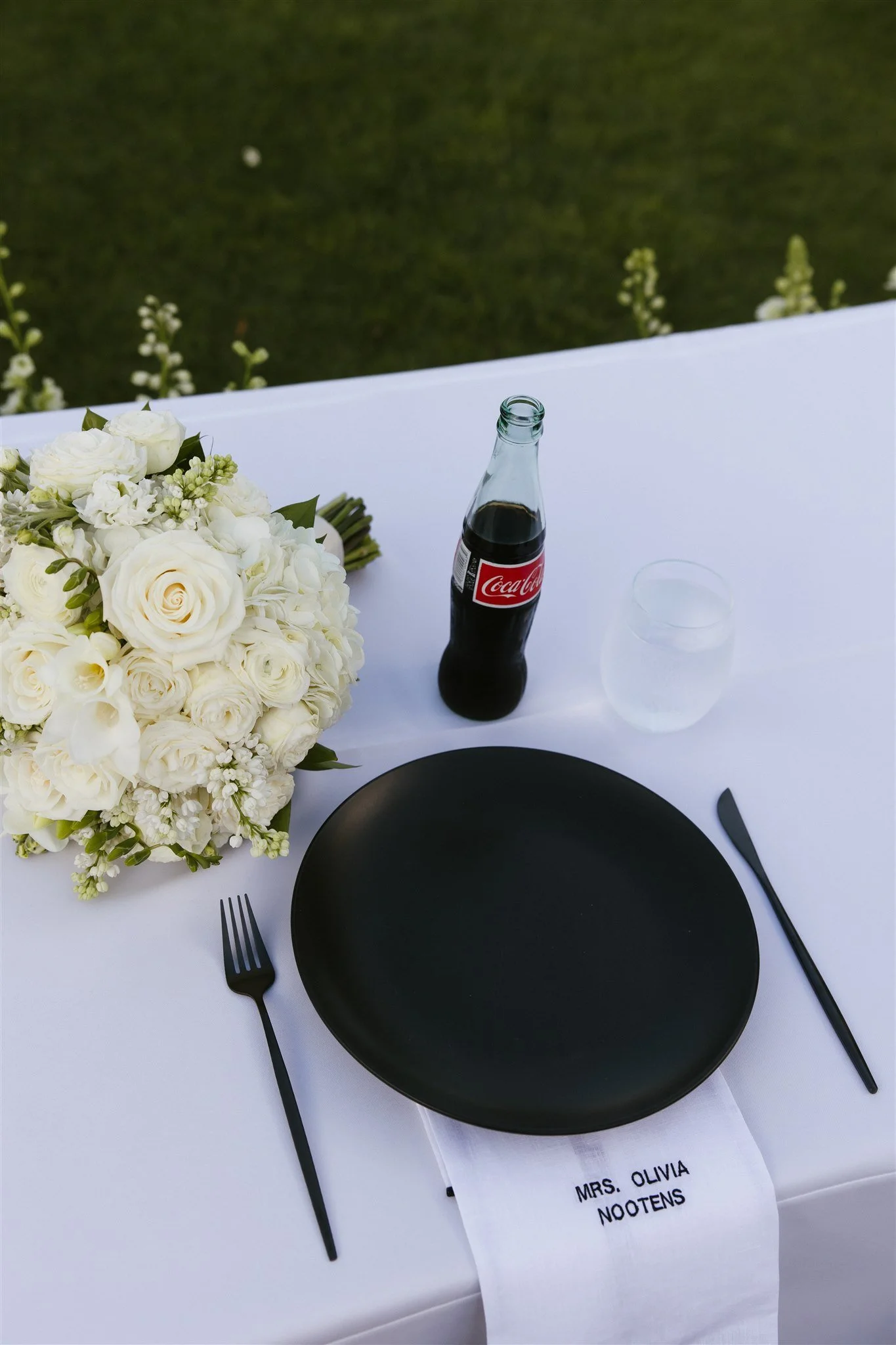 A table set for an event with a black plate, black cutlery, a bouquet of white flowers, a bottle of Coca-Cola, a glass of water, and a napkin with the name "Mrs. Olivia Nootens" on it.
