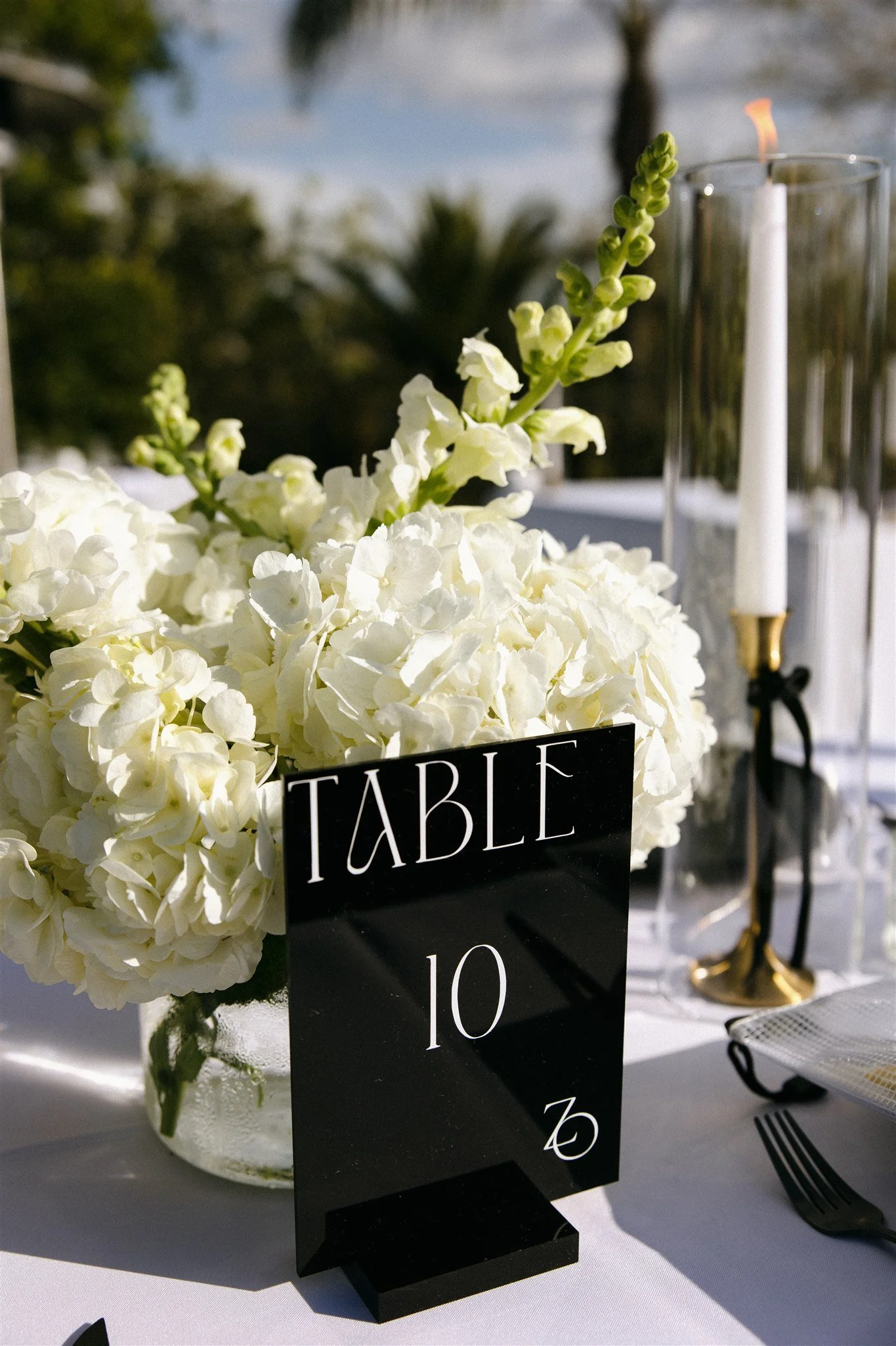Wedding table centerpiece with white hydrangeas, a black table number sign, and a candle in a glass holder outdoors.