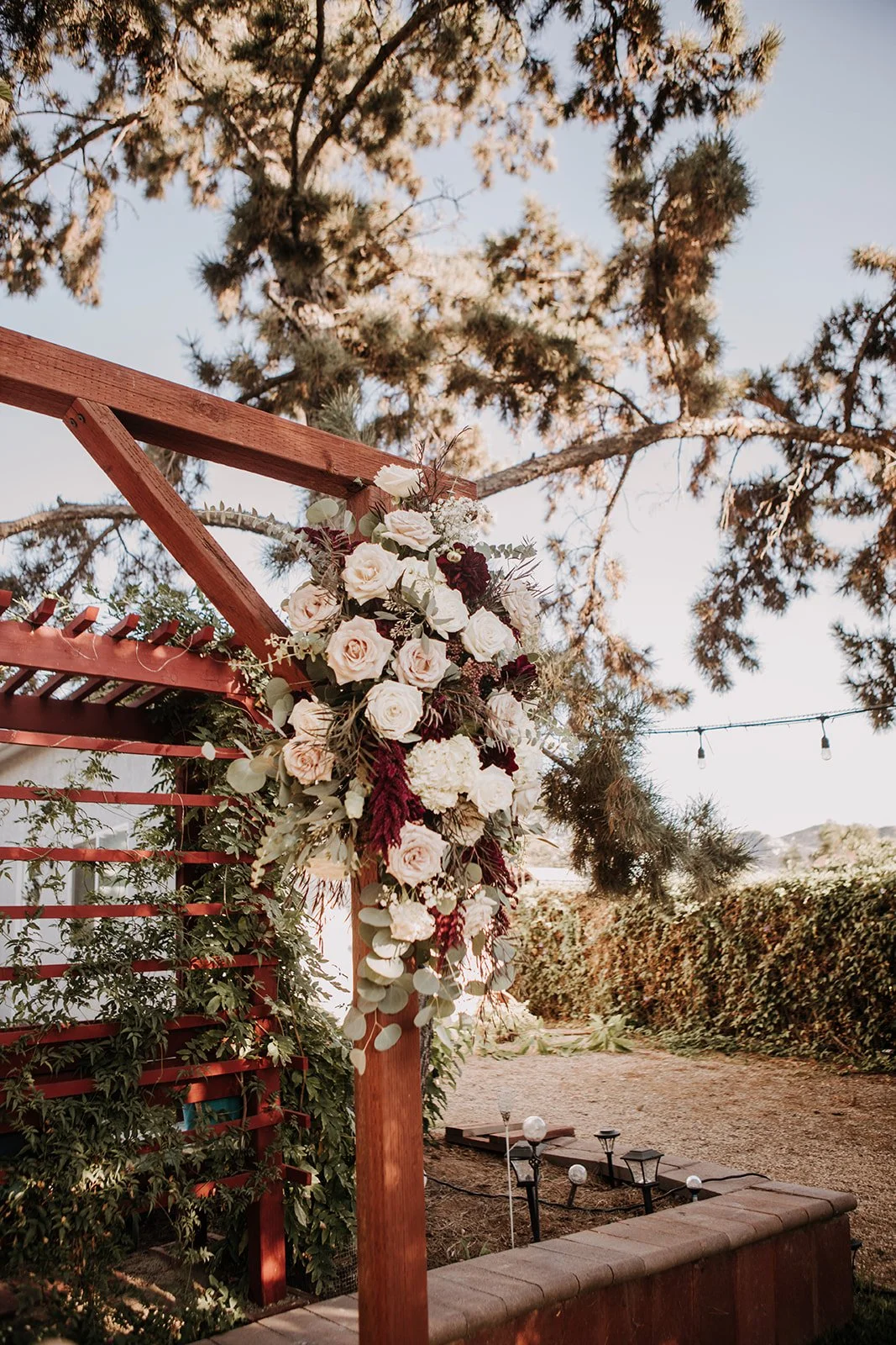 Flower arrangement with white and deep red roses, eucalyptus leaves, and other foliage attached to a wooden outdoor wedding arch.