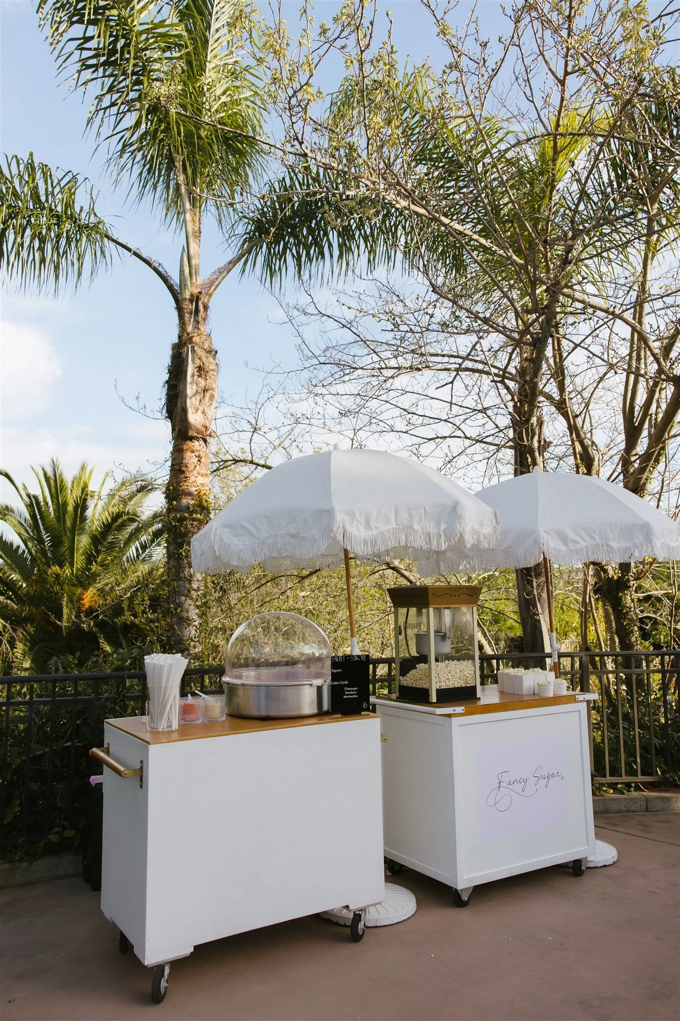 Food and drink stand with white umbrellas outdoors, featuring popcorn and cotton candy, surrounded by trees.