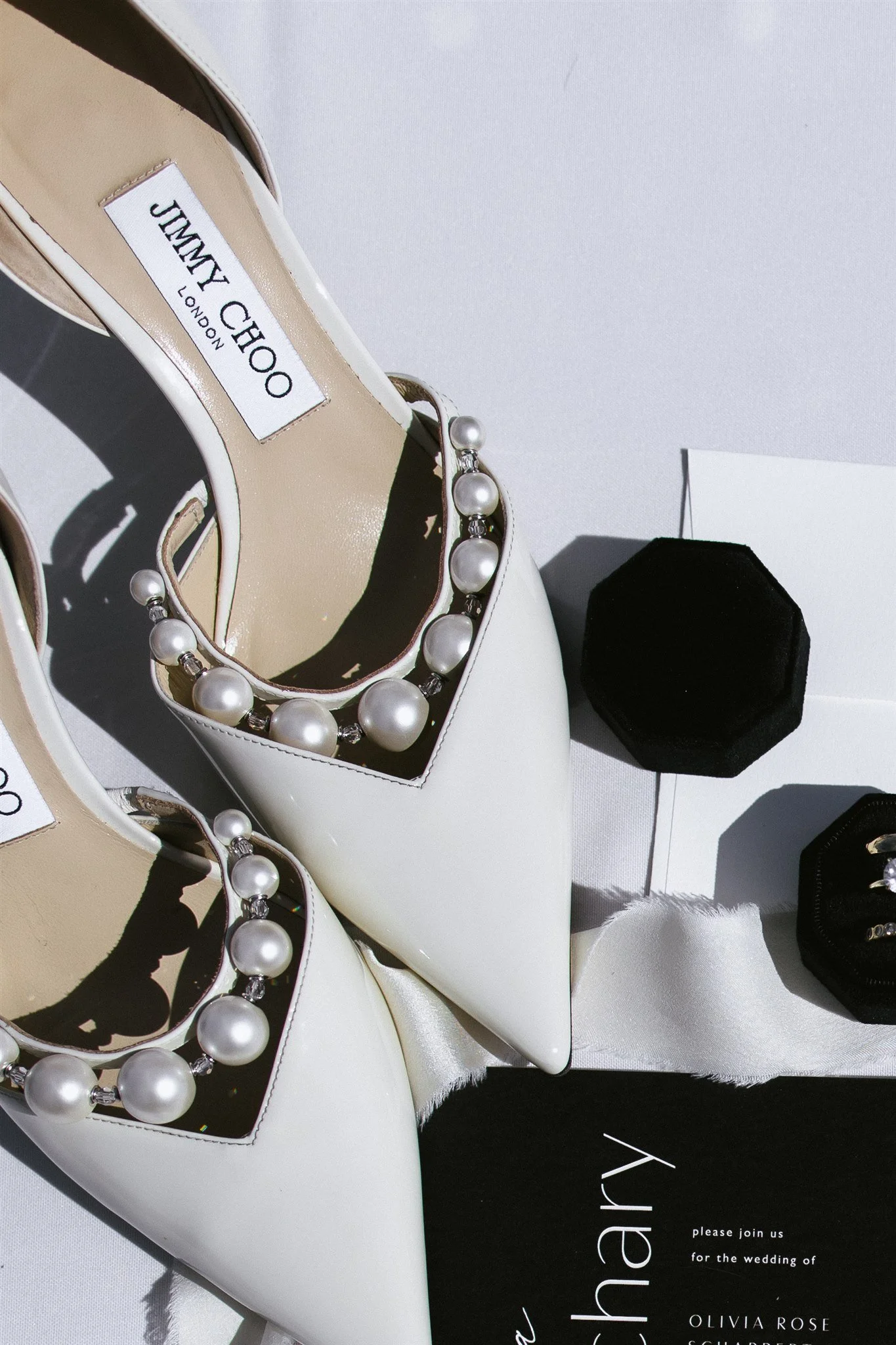 A pair of white Jimmy Choo high-heeled shoes with pearl necklaces inside the shoes, situated on a white surface. There are black and white jewelry boxes and an invitation card for a wedding on the surface.
