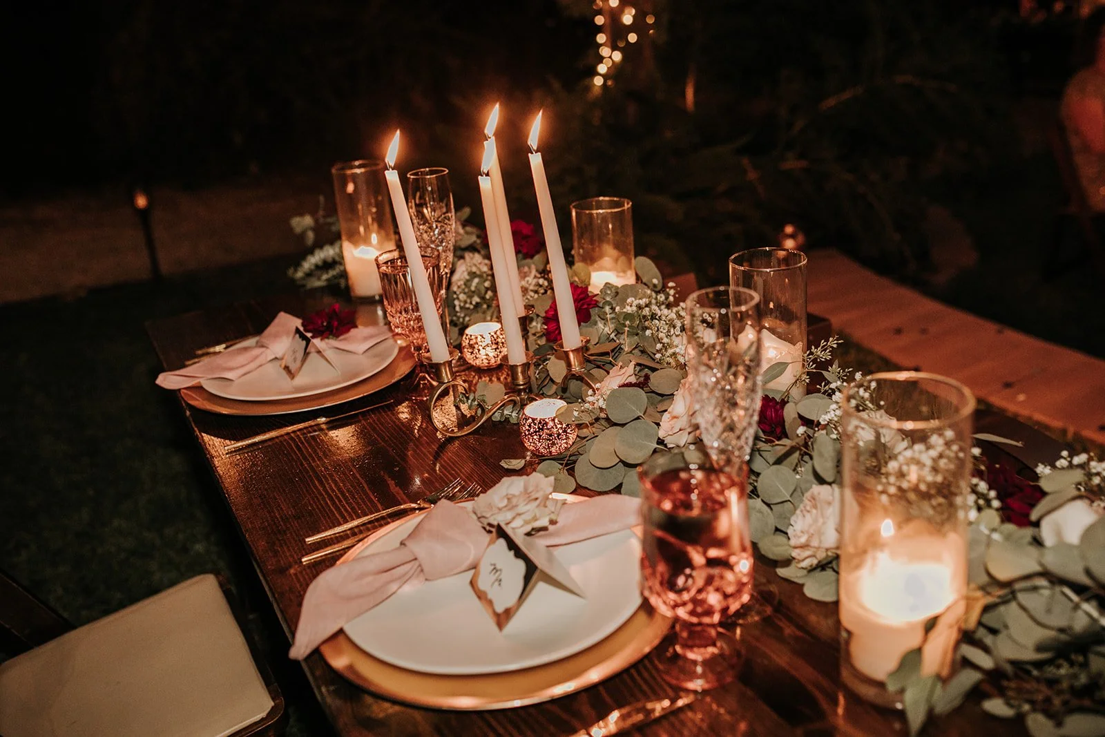 A candlelit dinner table set with floral arrangements, candles, and tableware for an event, possibly a wedding reception or romantic dinner.