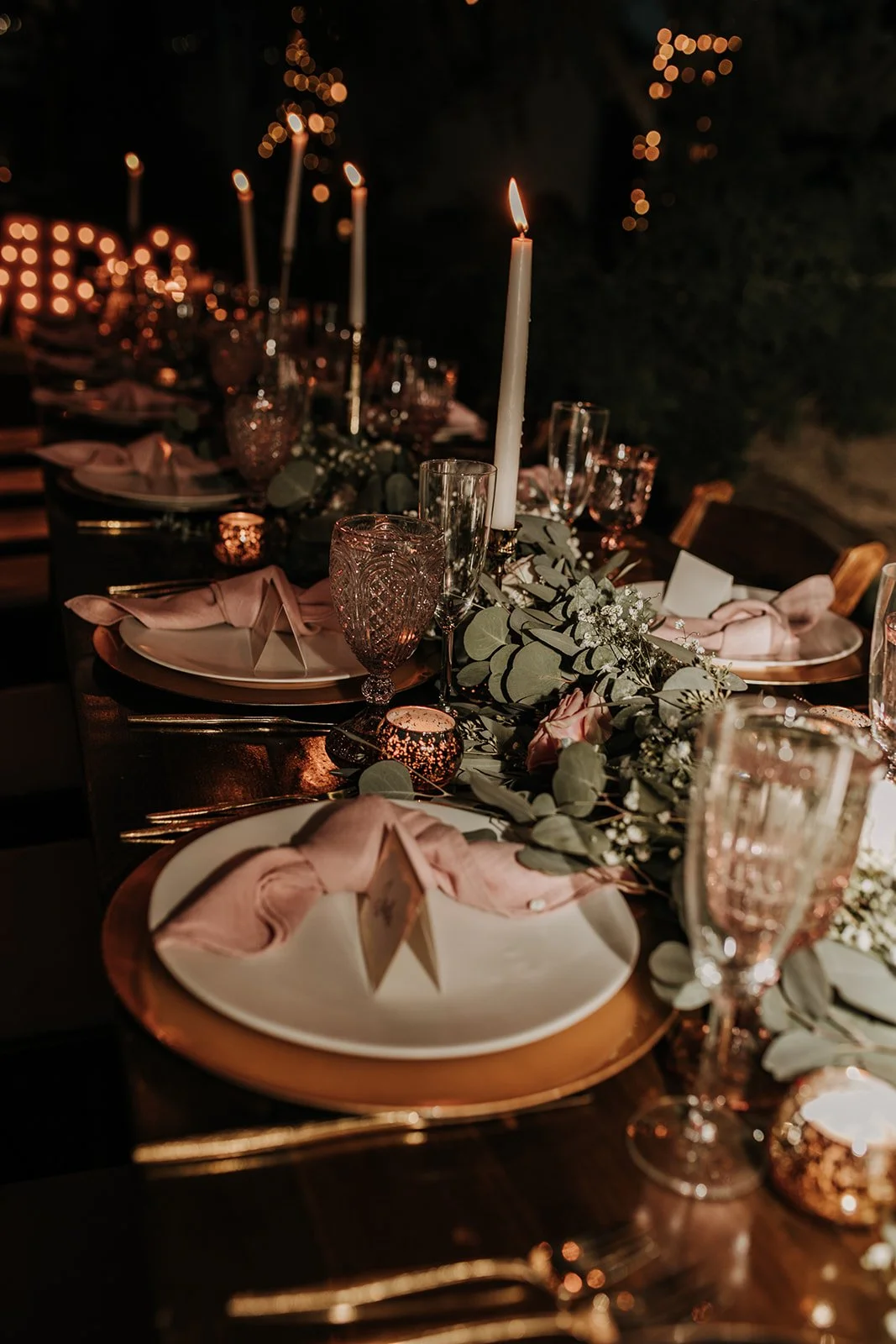 Elegant dinner table setting with white plates, pink napkins, glassware, a tall white candle, and a garland of greenery and flowers, illuminated by candlelight and string lights in a dark outdoor setting.