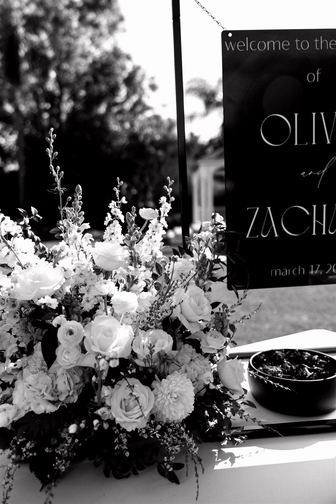 Black and white photo of a table with a large floral arrangement, a bowl of food, and a sign with text about an event at Olive and Zach on March 17.