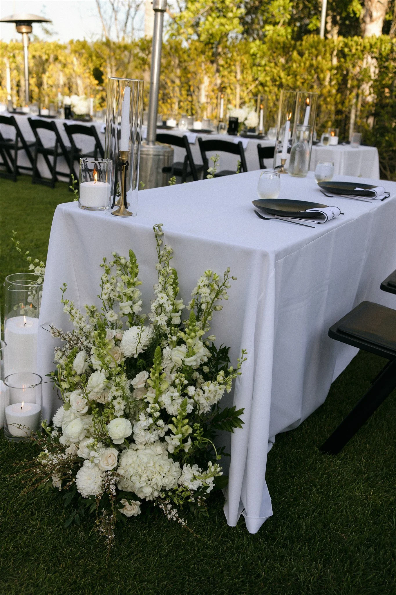 Elegant outdoor table setup with white tablecloth, black plates, white napkins, candles, vases, and a large white floral arrangement at the front.