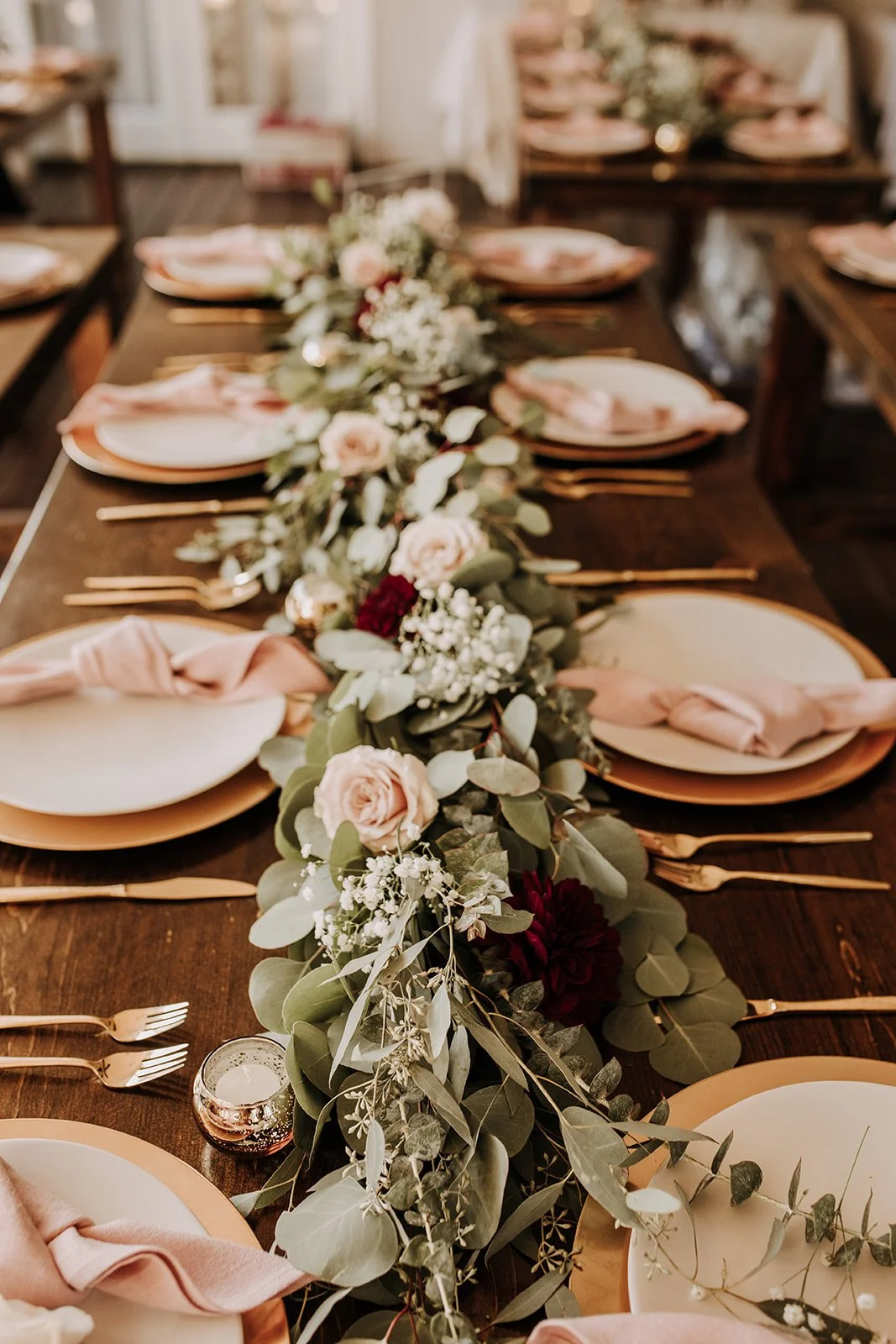 A decorated wedding or event table with a center floral runner consisting of pink roses, dark red flowers, eucalyptus and other greenery, gold cutlery, white and gold plates, pink napkins, and a small candle holder.