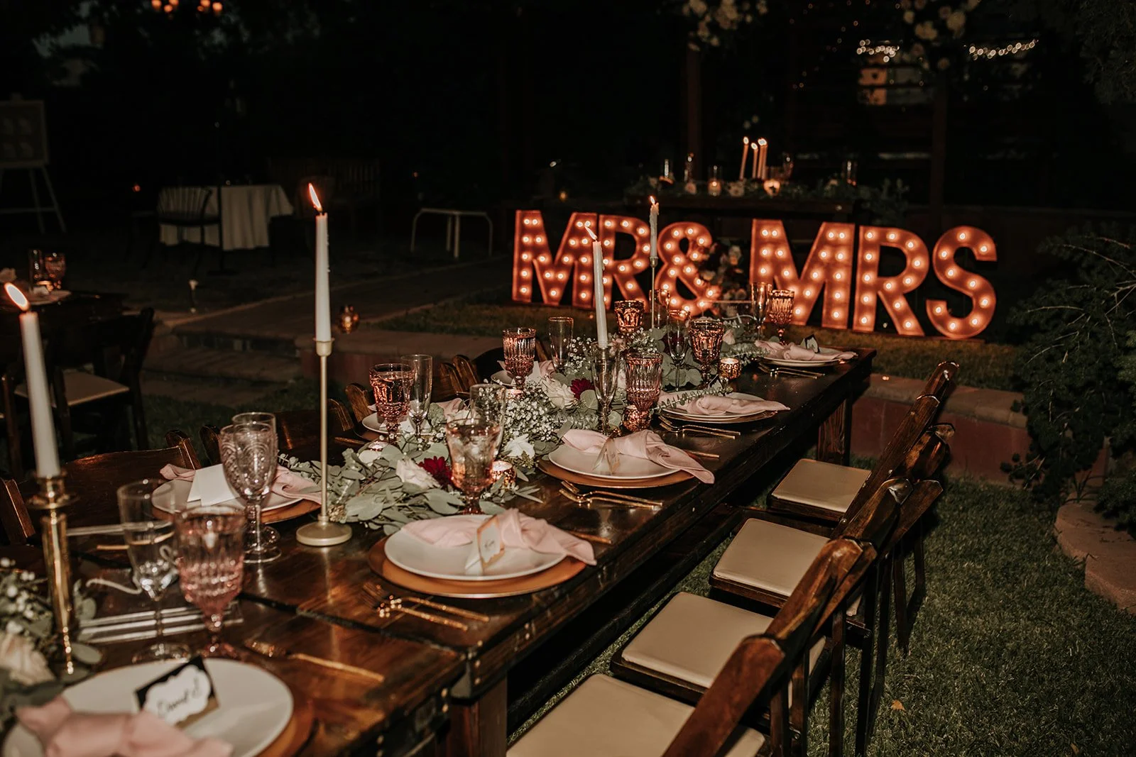 Outdoor wedding reception table decorated with candles, glassware, and floral centerpiece, with illuminated 'MR & MRS' sign in the background at night.