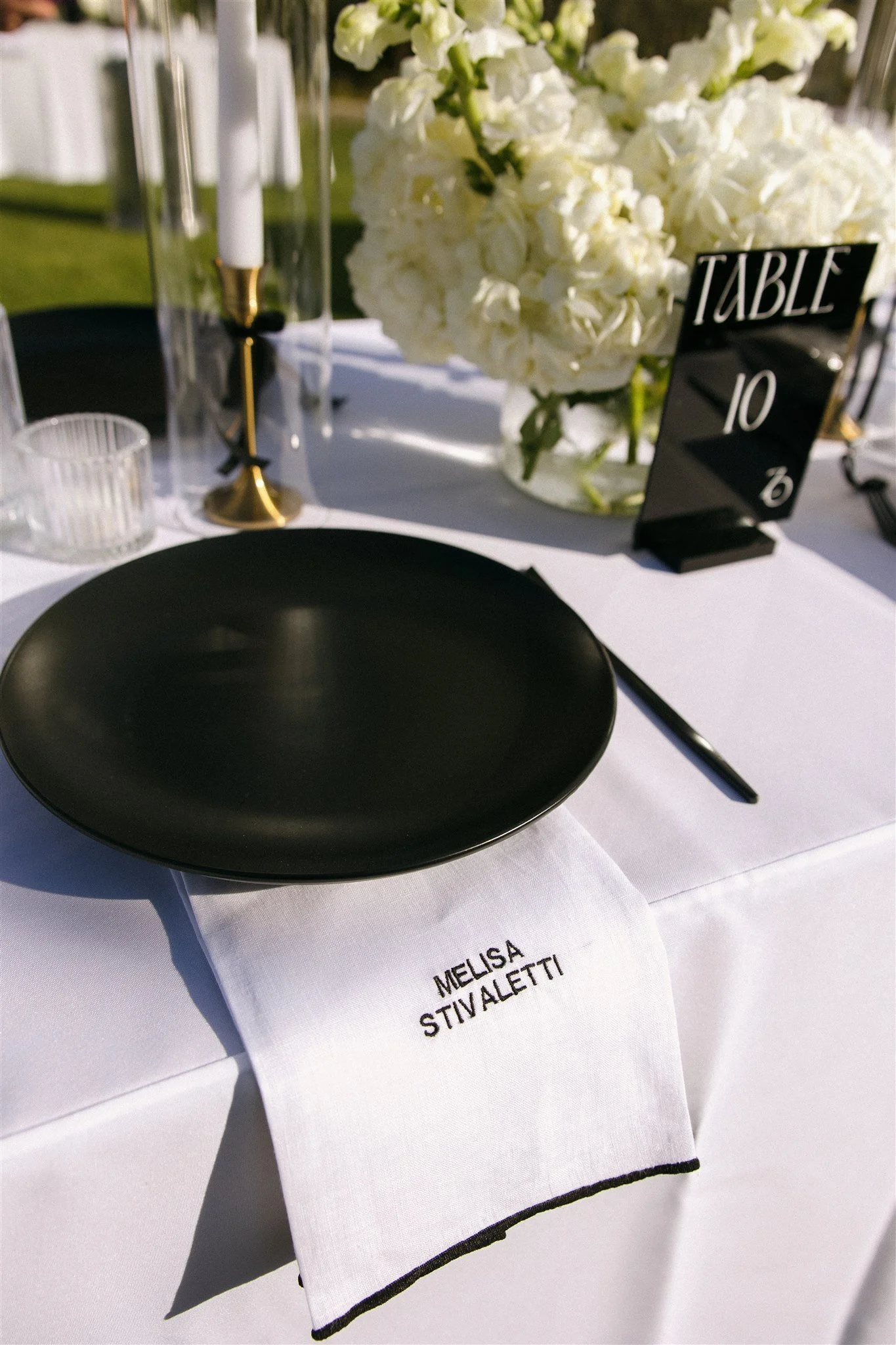 Elegant table setting with a black plate, a white cloth napkin embroidered with the name Melisa Stivaletti, a black table number sign, a glass vase with white hydrangeas, a black candlestick holder, and other table decor elements on a white tableclot