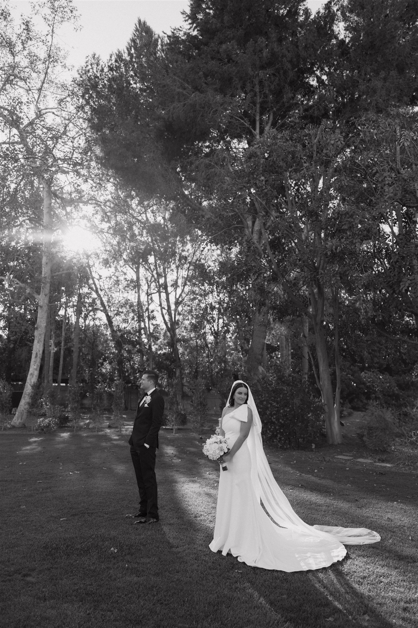 A bride in a white wedding dress and a groom in a black suit outdoors during sunset, with trees in the background.