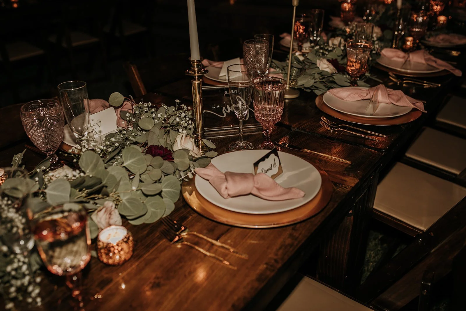 Elegant dinner table setup with a dark wooden surface, decorated with pink napkins, gold flatware, and floral centerpieces including eucalyptus, white roses, and baby's breath, illuminated by soft lighting and candles.