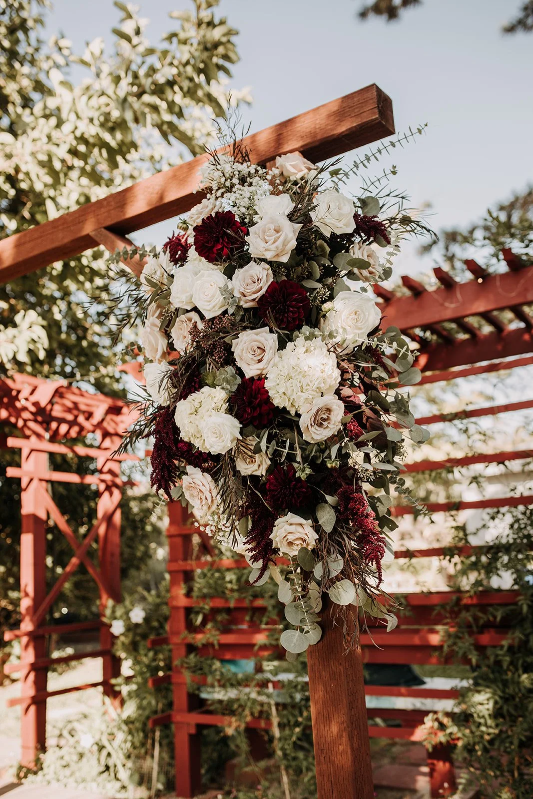 A floral arrangement with white roses, burgundy dahlias, and greenery hanging from a wooden arch at an outdoor wedding setting.