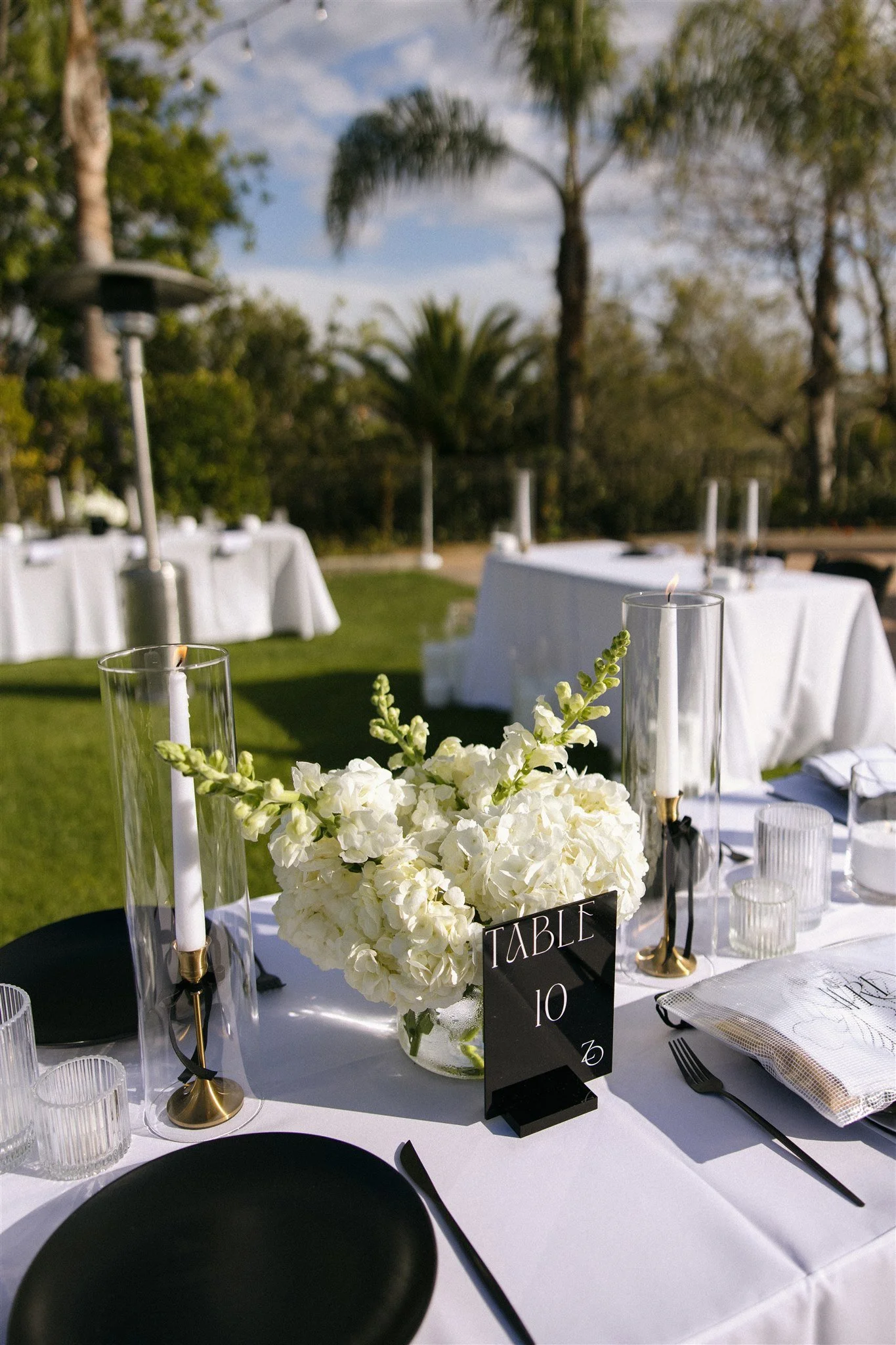Elegant outdoor event table setup with white flowers, candles in glass holders, and place settings on a white tablecloth, surrounded by greenery and palm trees.