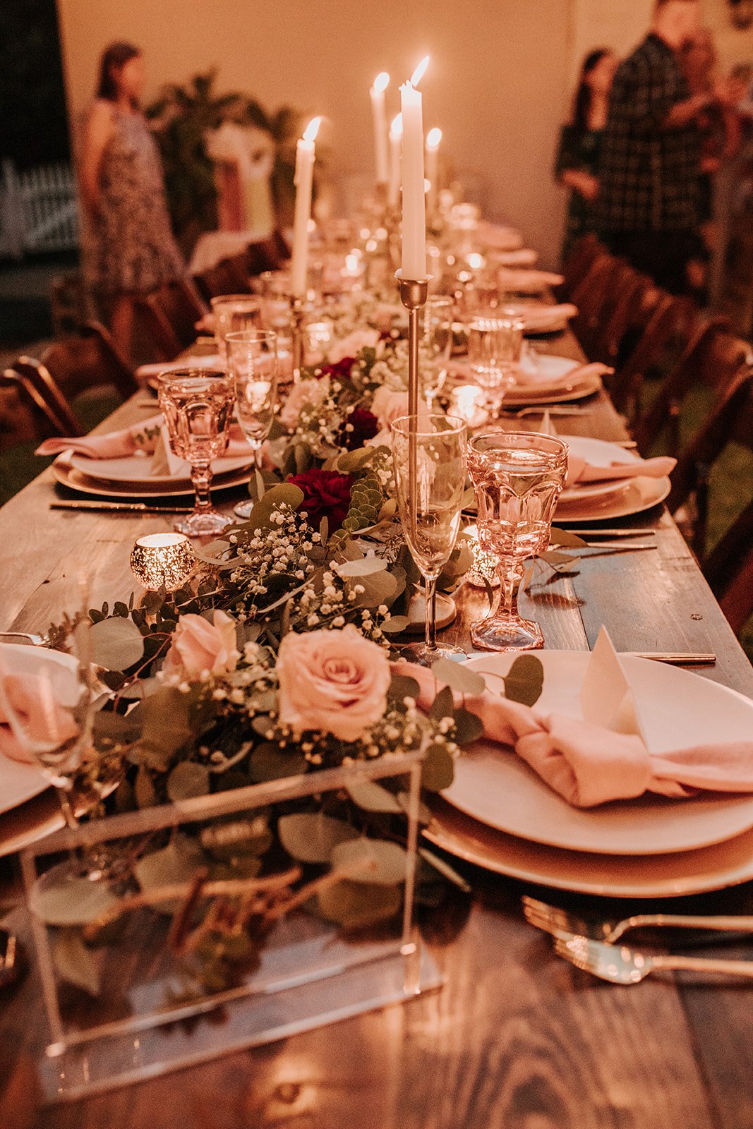 A long dining table decorated with pink roses, baby's breath, greenery, candles, and elegant glassware, set for a formal event or celebration.