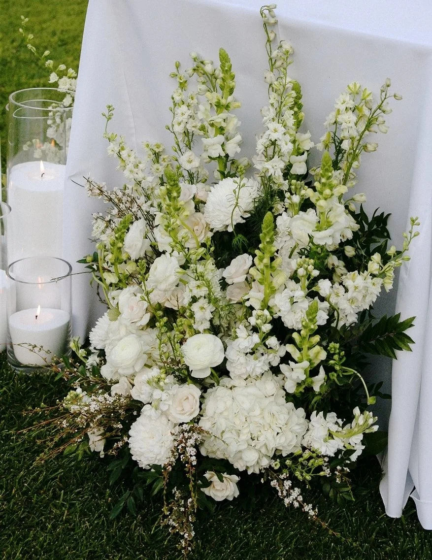 A white floral arrangement with white flowers and greenery, placed on grass next to lit candles and white drapes, likely for a funeral or memorial service.