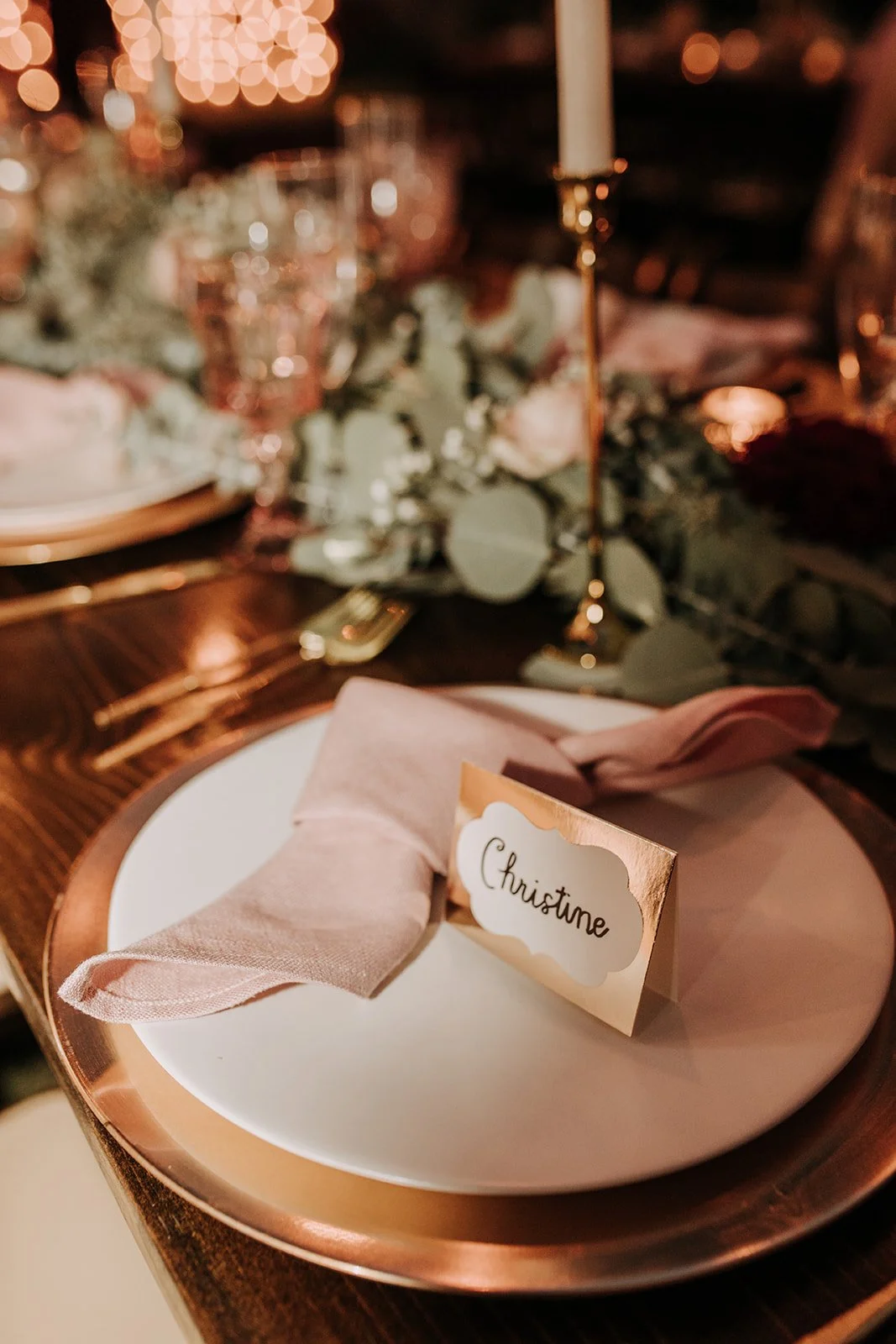 A place setting on a dinner table with a white plate, pink napkin, and a gold place card labeled "Christine." The table is decorated with a floral centerpiece and gold accents, and is illuminated with warm lighting and candlelight.