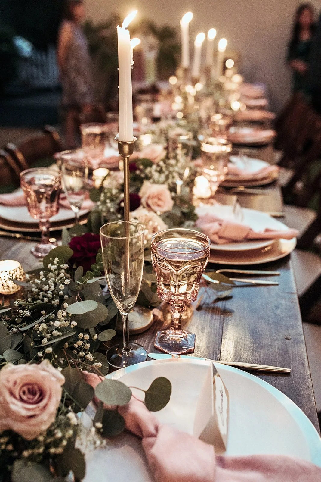 A beautifully set dinner table with pink and white floral arrangements, candles, pink wine glasses, and place settings with napkins, with soft candlelight illuminating the scene.