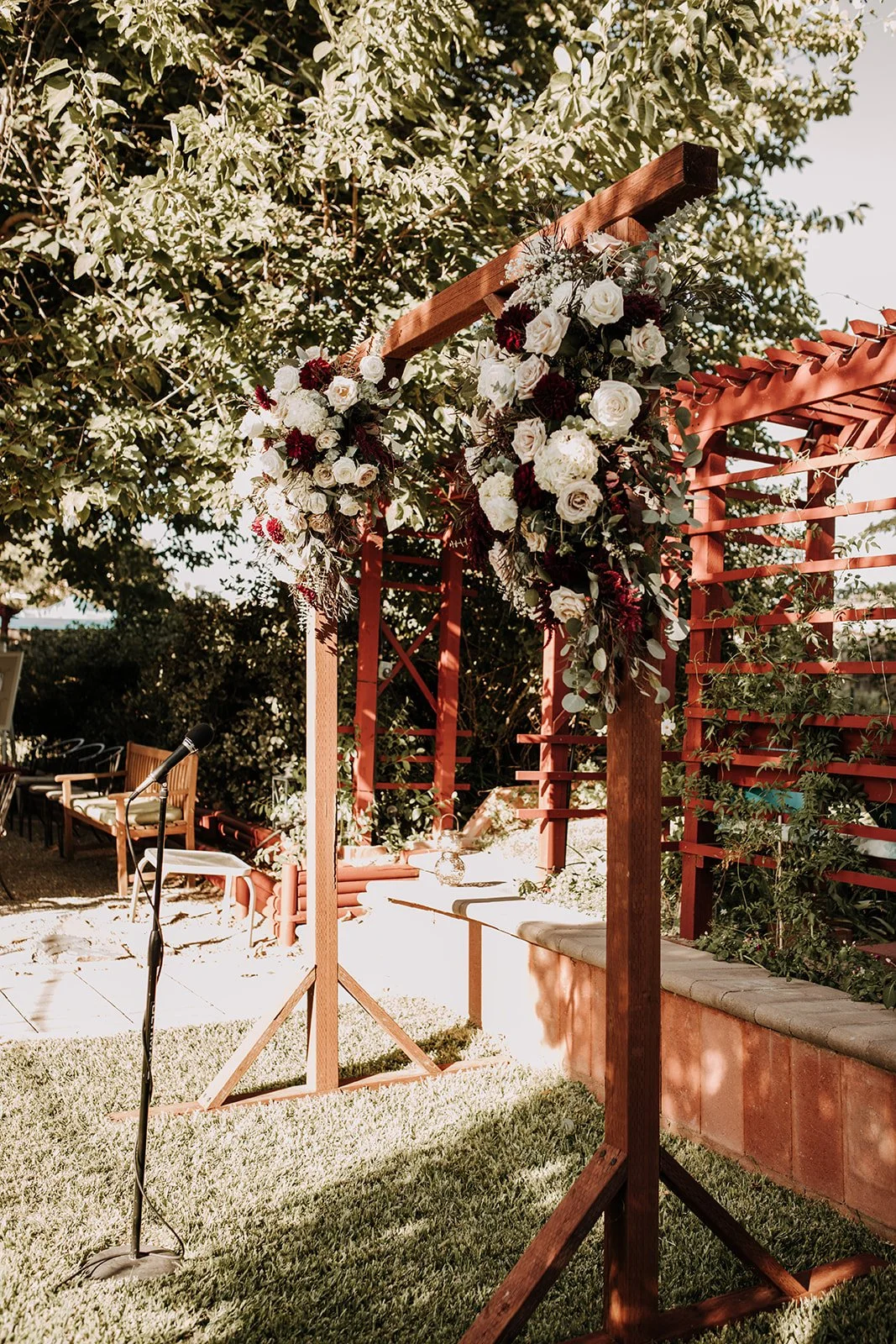 Outdoor wedding or event setup with a wooden arch decorated with white and dark red flowers, greenery, and draped fabric, situated on a grassy area under a large tree.