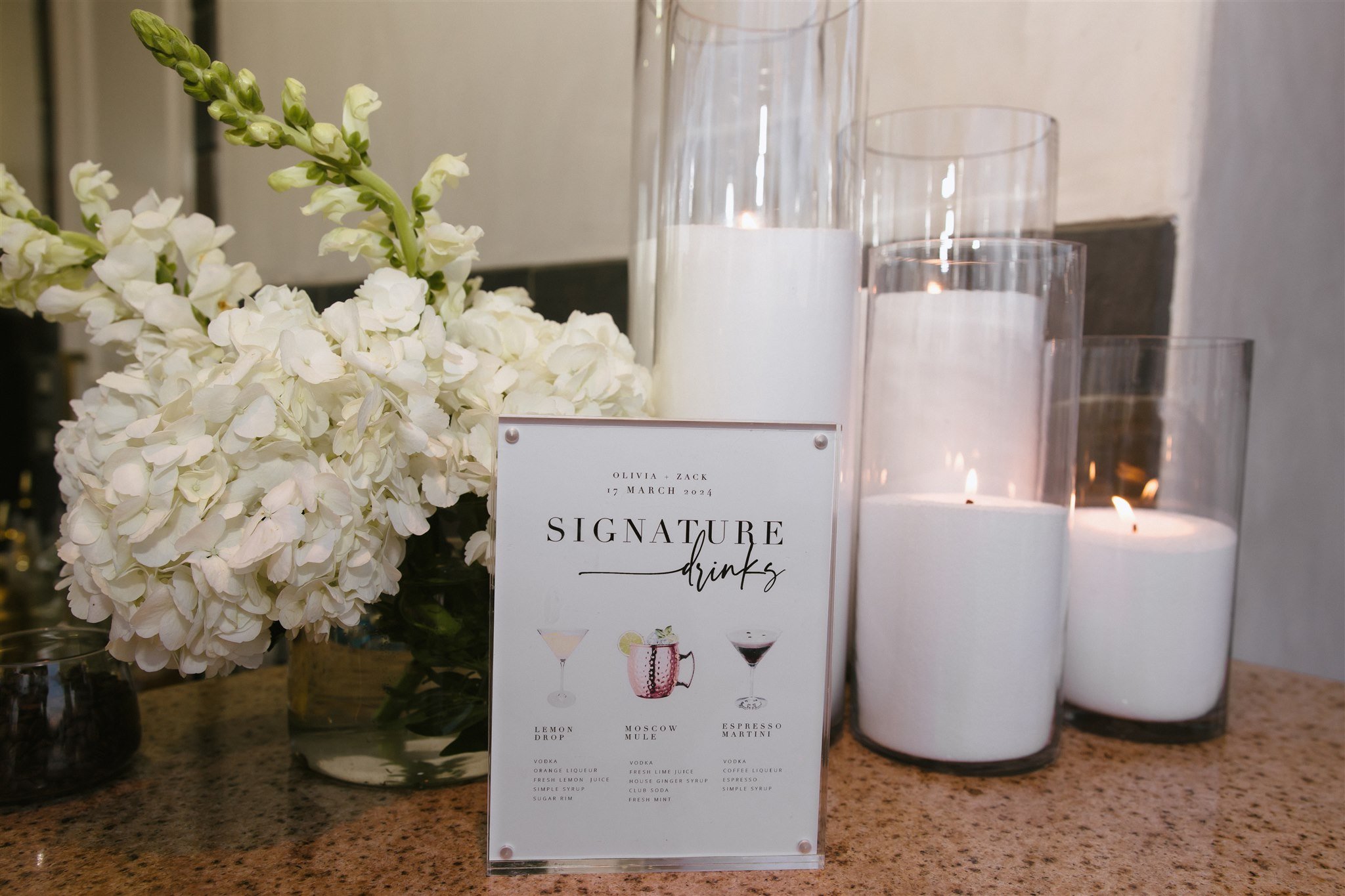 A floral arrangement of white hydrangeas, four tall white candles in glass holders with lit wicks, and a printed menu stand displaying signature drinks on a brown granite countertop.