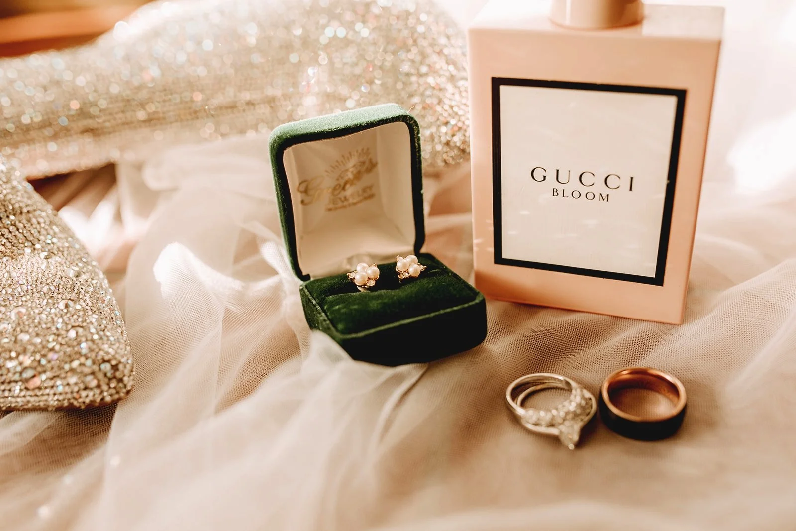 A pair of pearl earrings in a green velvet jewelry box, a Gucci Bloom perfume box, and three rings on silky fabric surrounded by a glittery clutch.