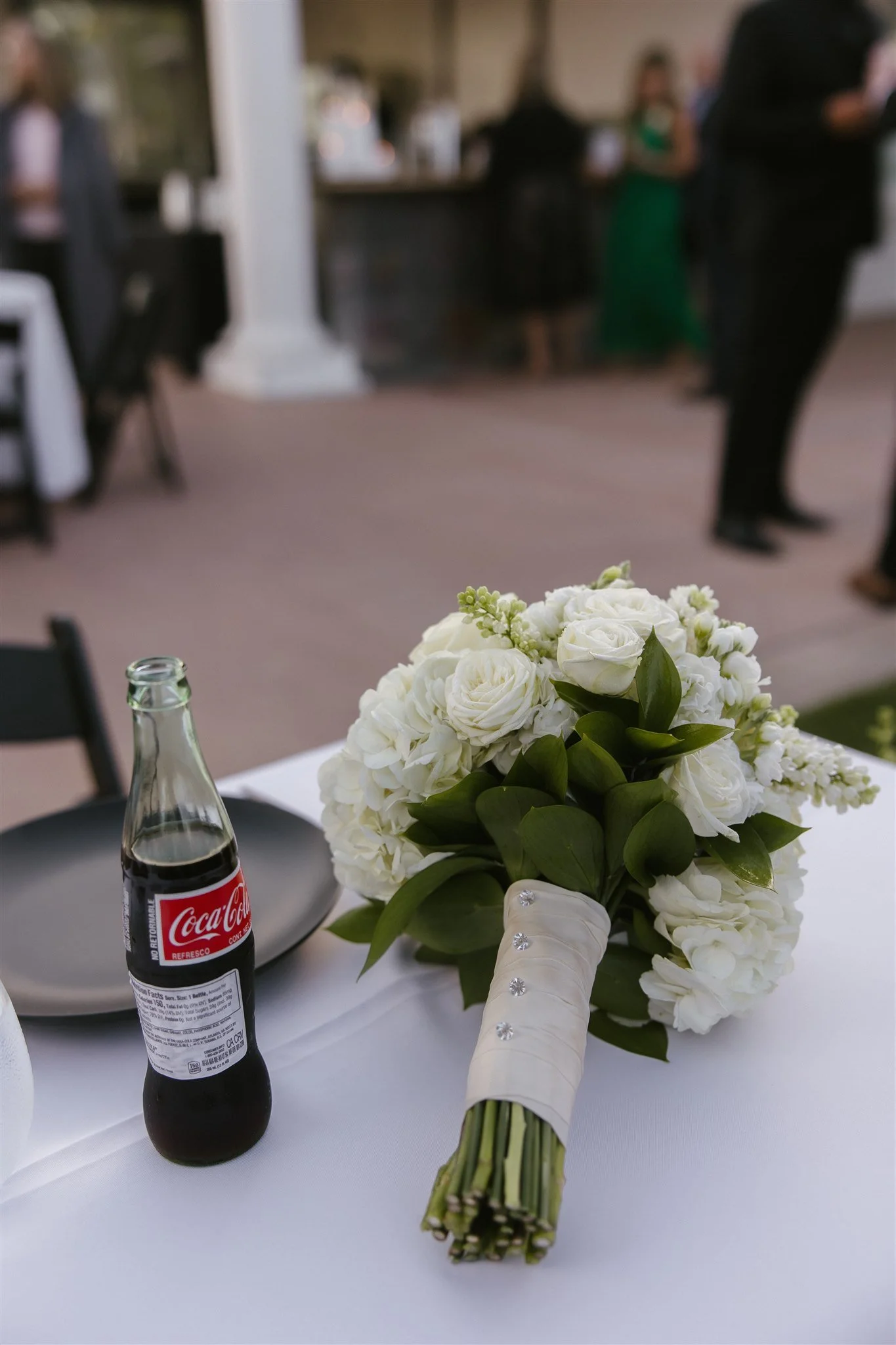 A wedding bouquet of white roses and hydrangeas with green leaves, placed on a white tablecloth next to a small, open Coke bottle, with a background of a blurred outdoor event or reception.