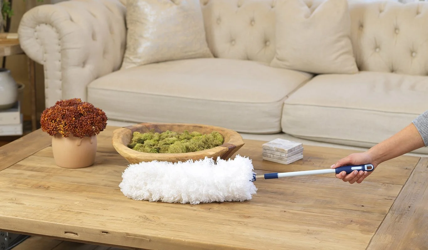 Person vacuuming a wooden coffee table with decorative items including a potted plant, a bowl of moss, and a stack of coasters, in a living room with a cream-colored sofa and an armchair.