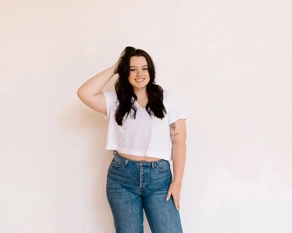 A young woman with long dark hair wearing a white cropped t-shirt and blue jeans, smiling and standing against a plain light-colored wall.