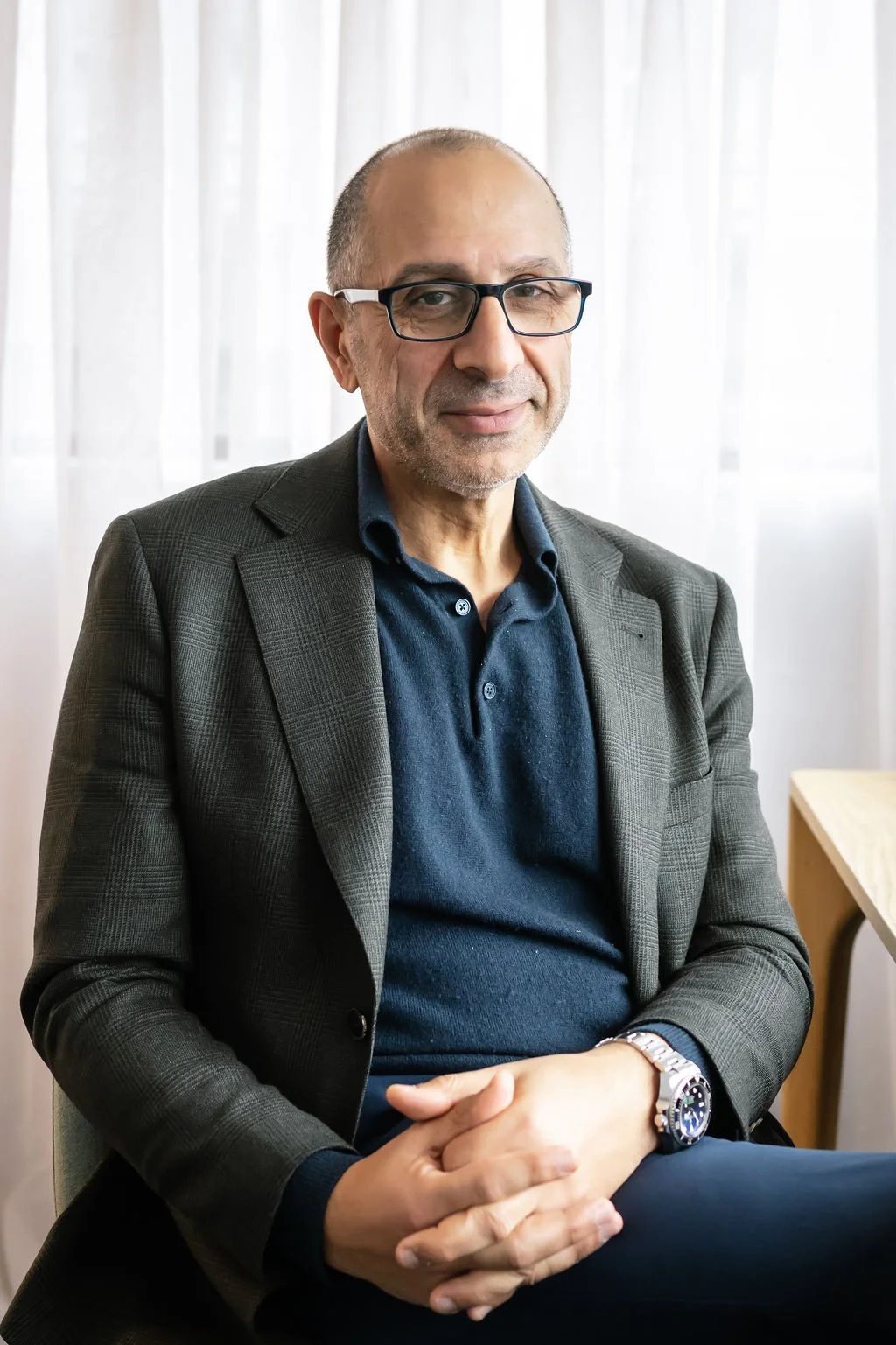 A middle-aged man with glasses, wearing a dark blue polo shirt and gray blazer, sitting indoors near a window with sheer white curtains.