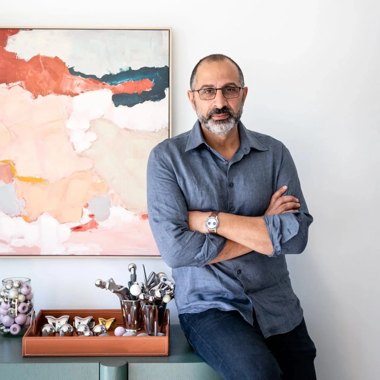 Dr Elie Khoury Orthopaedic Surgeon - A man with glasses and a beard, wearing a blue shirt, standing with arms crossed in front of a colorful abstract painting. There are decorative ornaments on a tray and a jar on a green table.