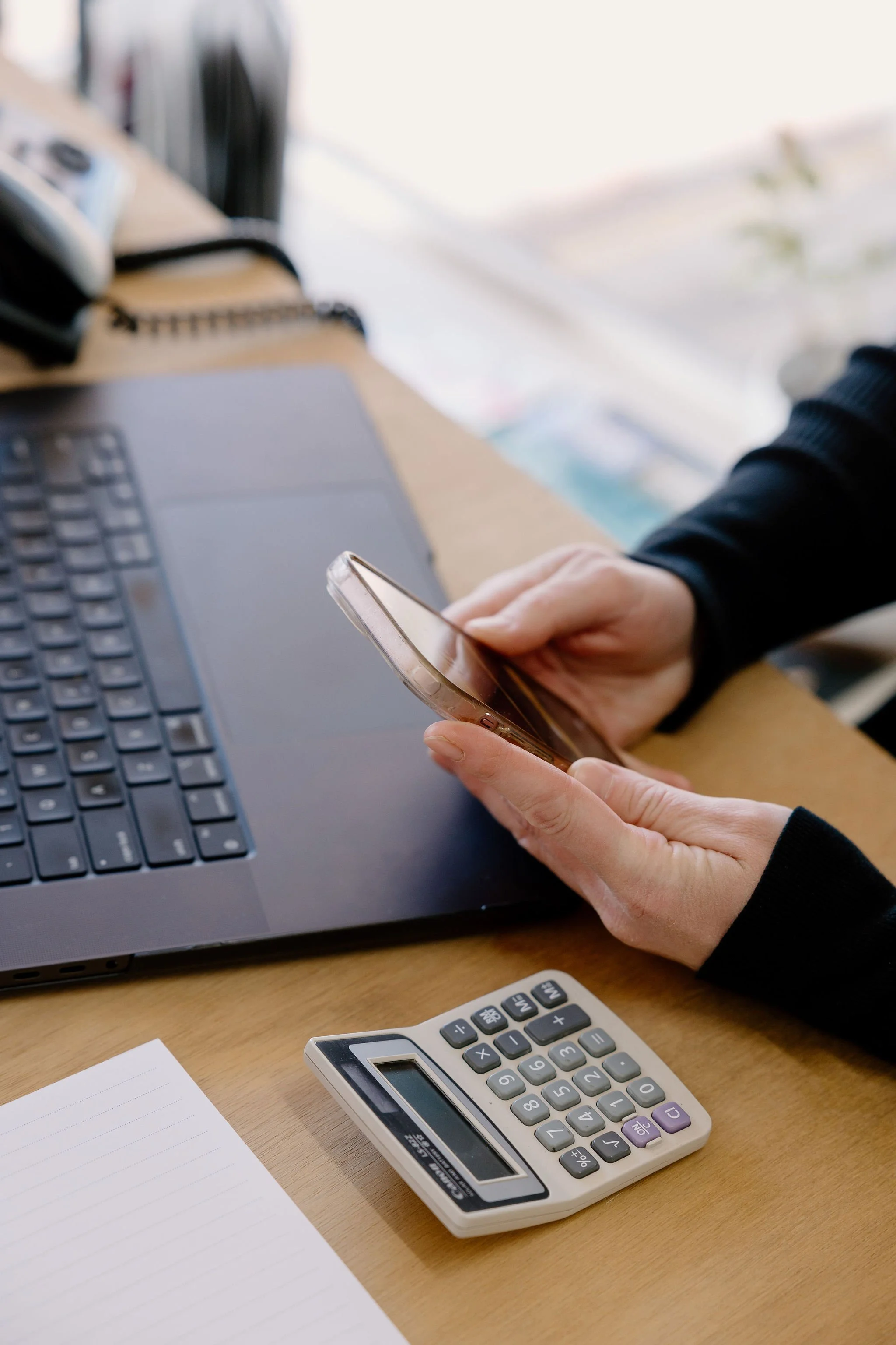 Person using a smartphone at a desk with a laptop, calculator, and notepad.