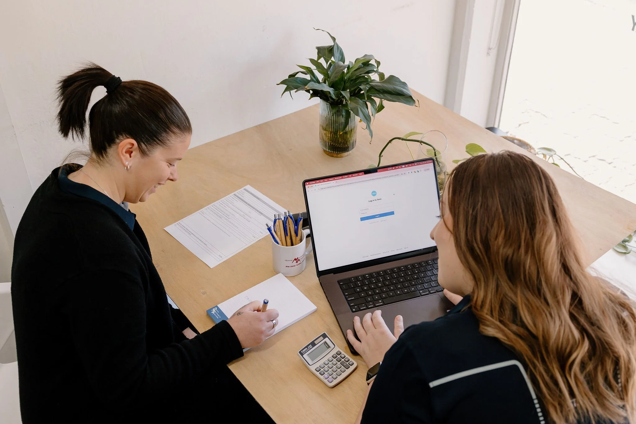 Two women sitting at a wooden table in a bright room, working together with a laptop, notebooks, a calculator, and pens, with a large plant on the table.
