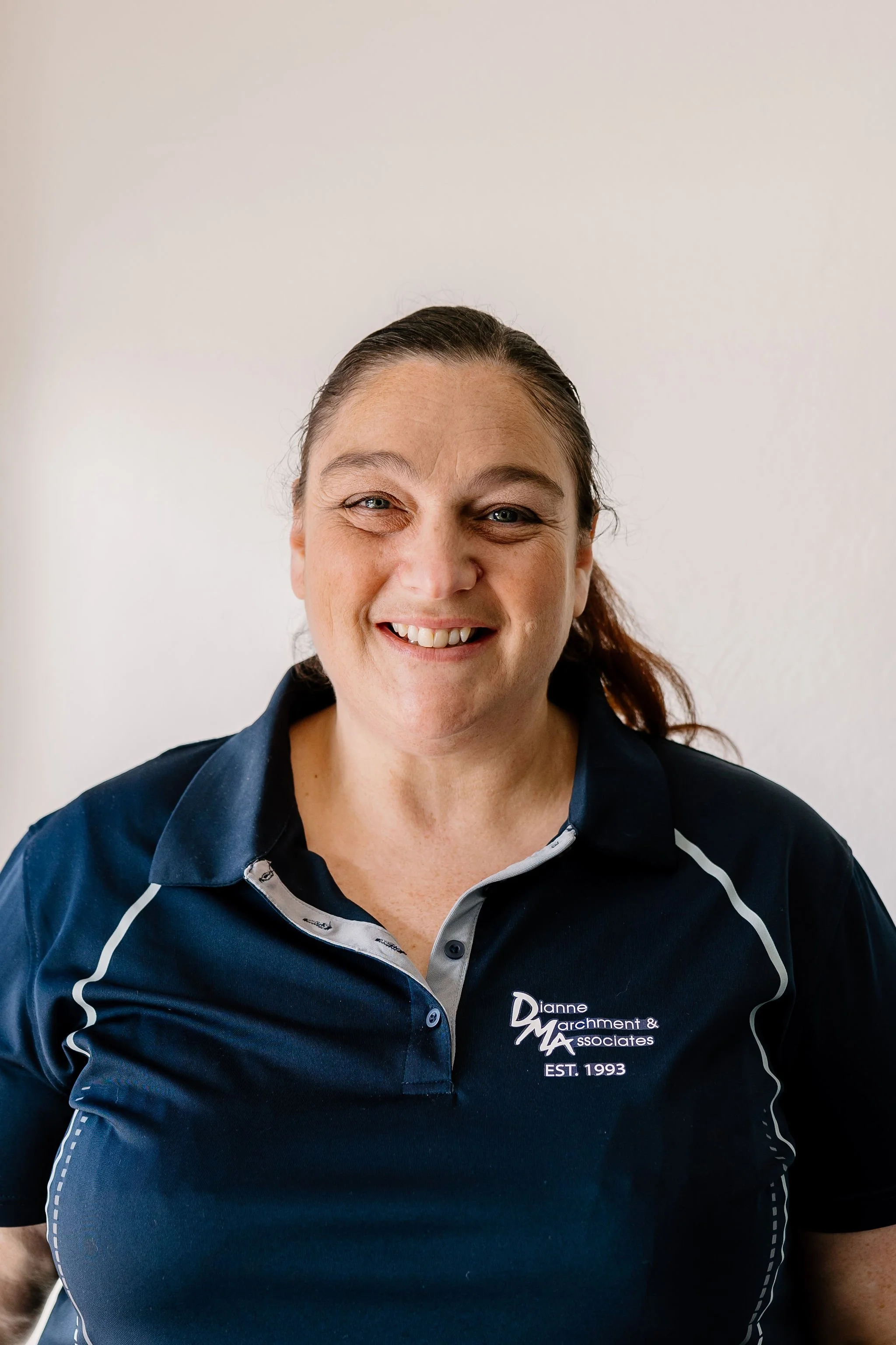 Alison smiling, wearing a navy blue polo shirt with embroidered text 'Dianne Marchment & Associates' and 'EST. 1993' on the front, standing in front of a plain off-white wall.