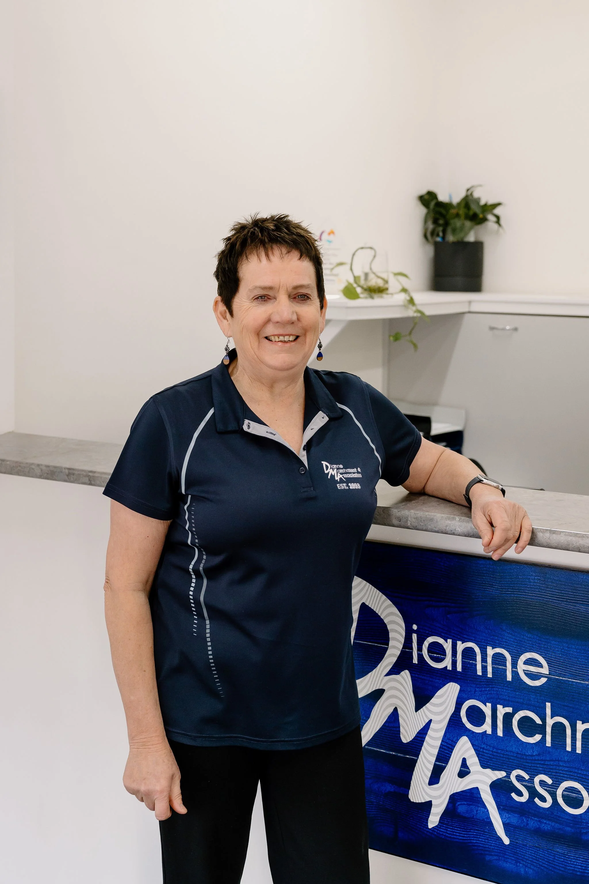 Small business owner Dianne Marchment, in a navy blue polo shirt, standing at the reception counter with a sign that says Dianne Marchment and Associates.