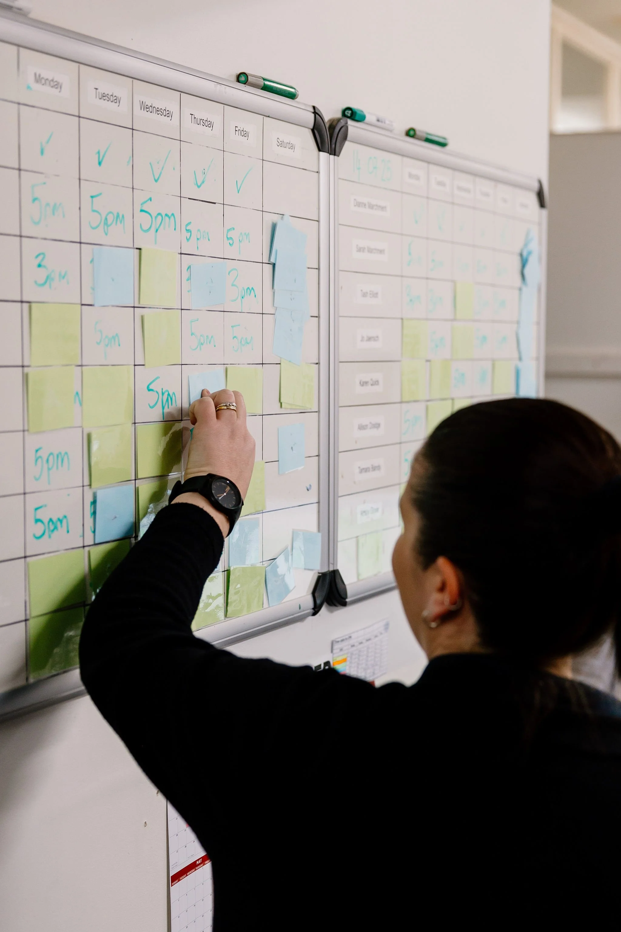 A woman organizing work schedules on a whiteboard using sticky notes in an office setting.
