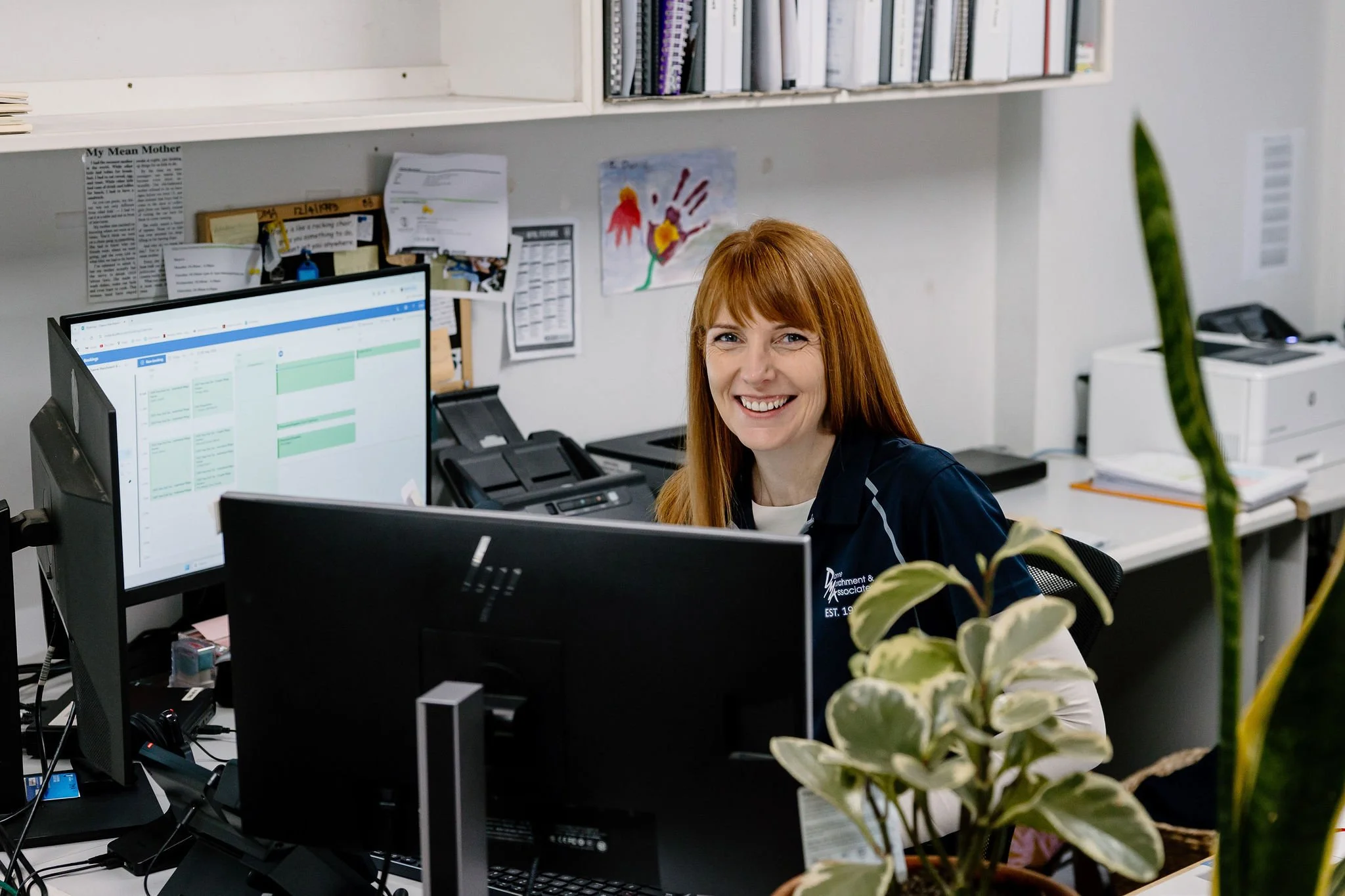A woman with red hair smiling at her computer in an office setting with dual monitors, a plant in the foreground, and office supplies on the desk.