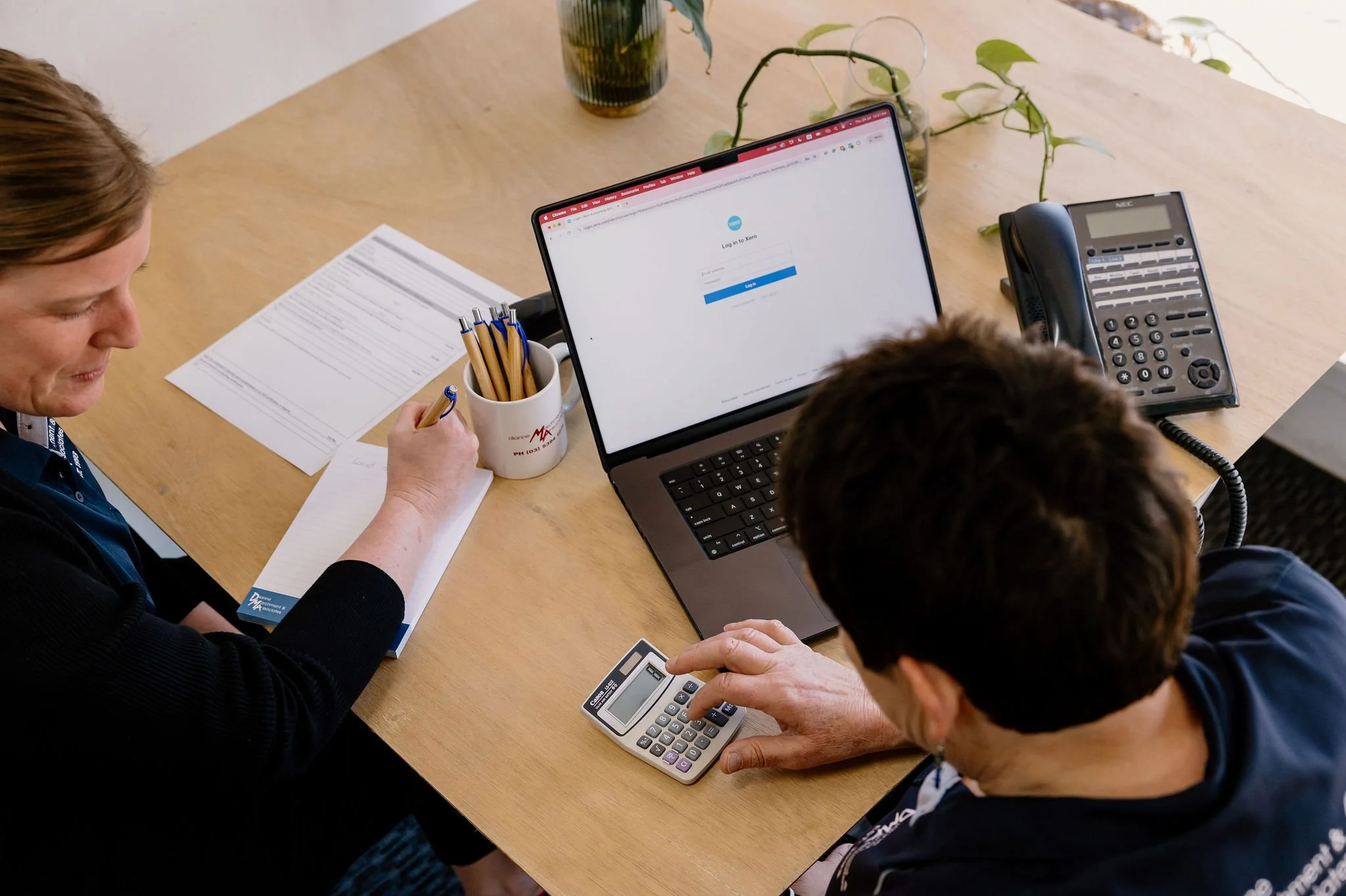 Di and Tash working together. The woman has a notepad and pen; the other is using a calculator. A laptop displays a login page. The desk holds a landline phone, a pot with pens, documents, and a plant.