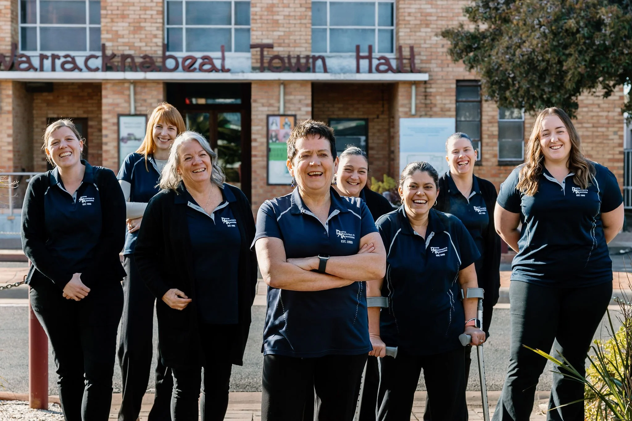 Di Marchment and Associates team in navy blue polo shirts standing in front of the Warracknabeal Town Hall, outside on the sidewalk during daytime.