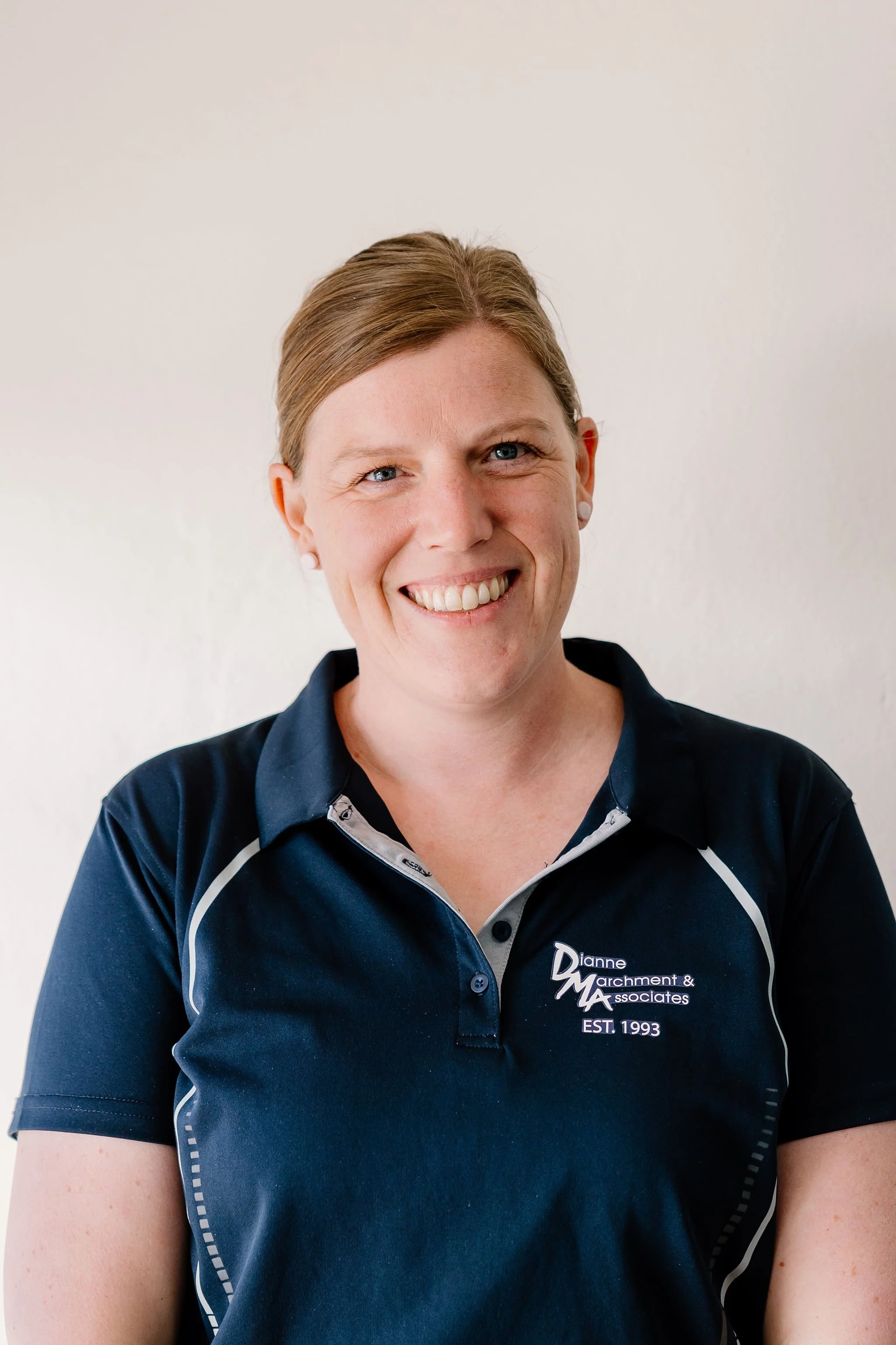 Kristy woman standing against a plain wall, wearing a navy blue polo shirt with branding on the chest, smiling at the camera.