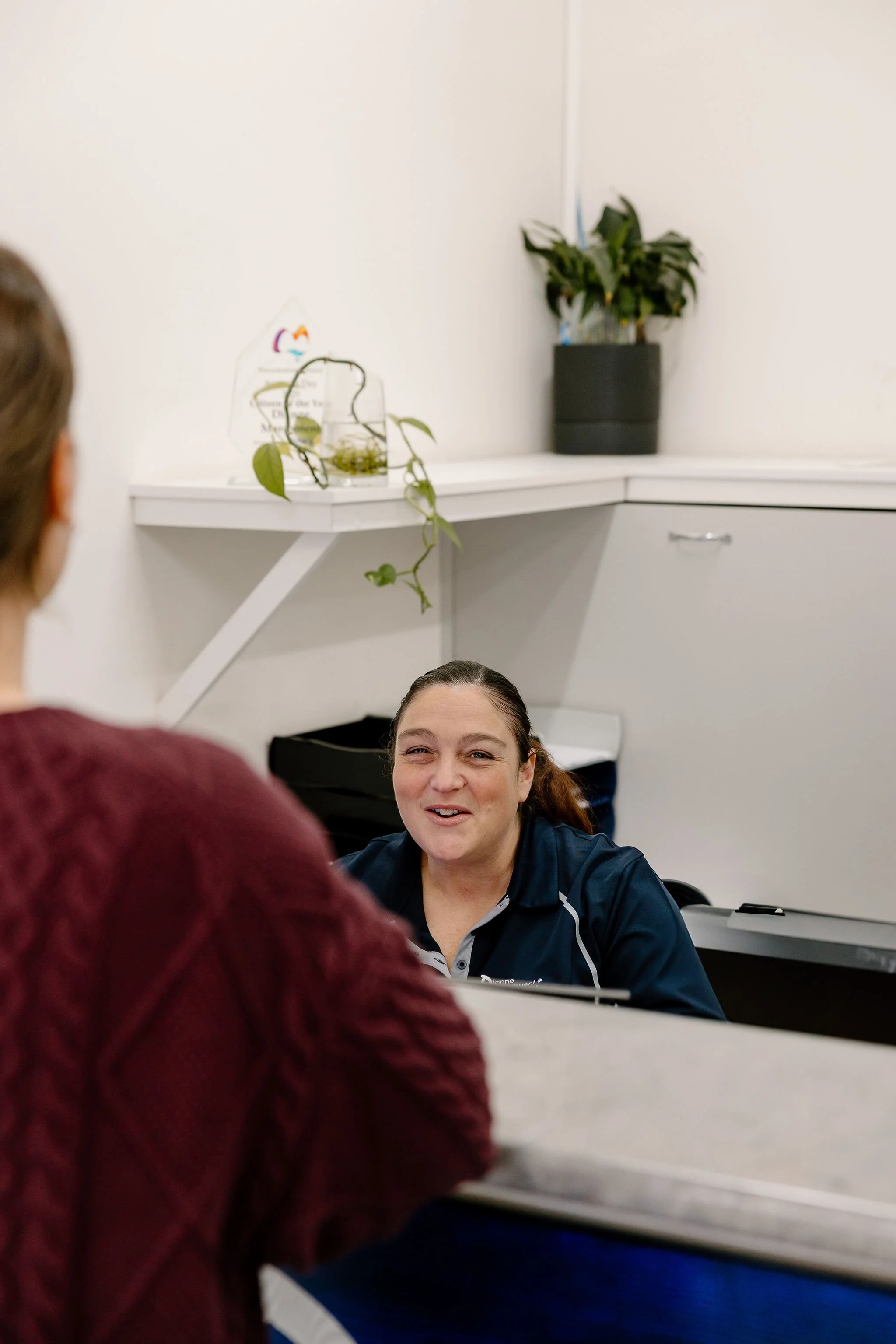 A woman with dark hair talking to another woman with brown hair in a white office, with plants and decor in the background.