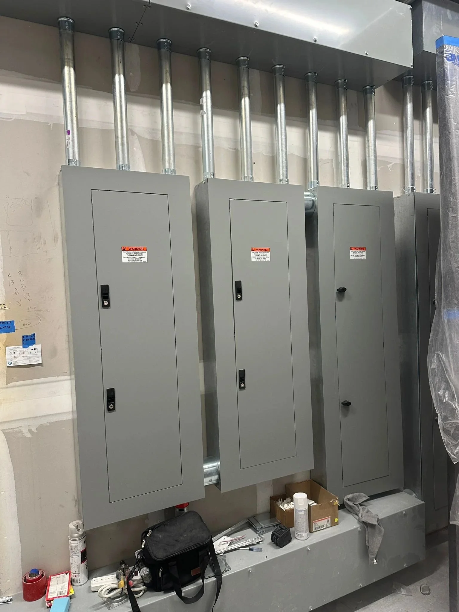Three electrical panels with metal conduit pipes above, mounted on a wall in an unfinished room, with tools and supplies on the floor below.
